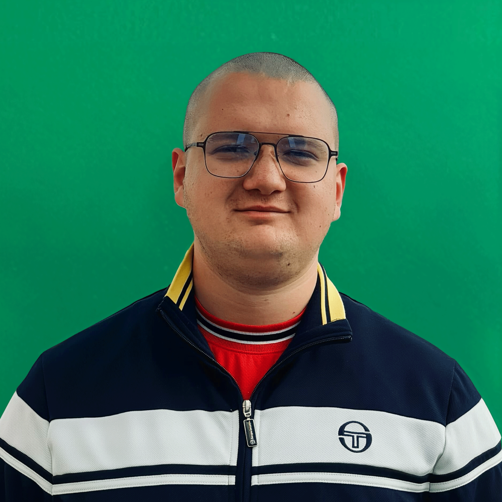 Portrait of a man with glasses, shaved head, wearing a black and white zip-up jacket with yellow collar and a red shirt underneath, against a green background.