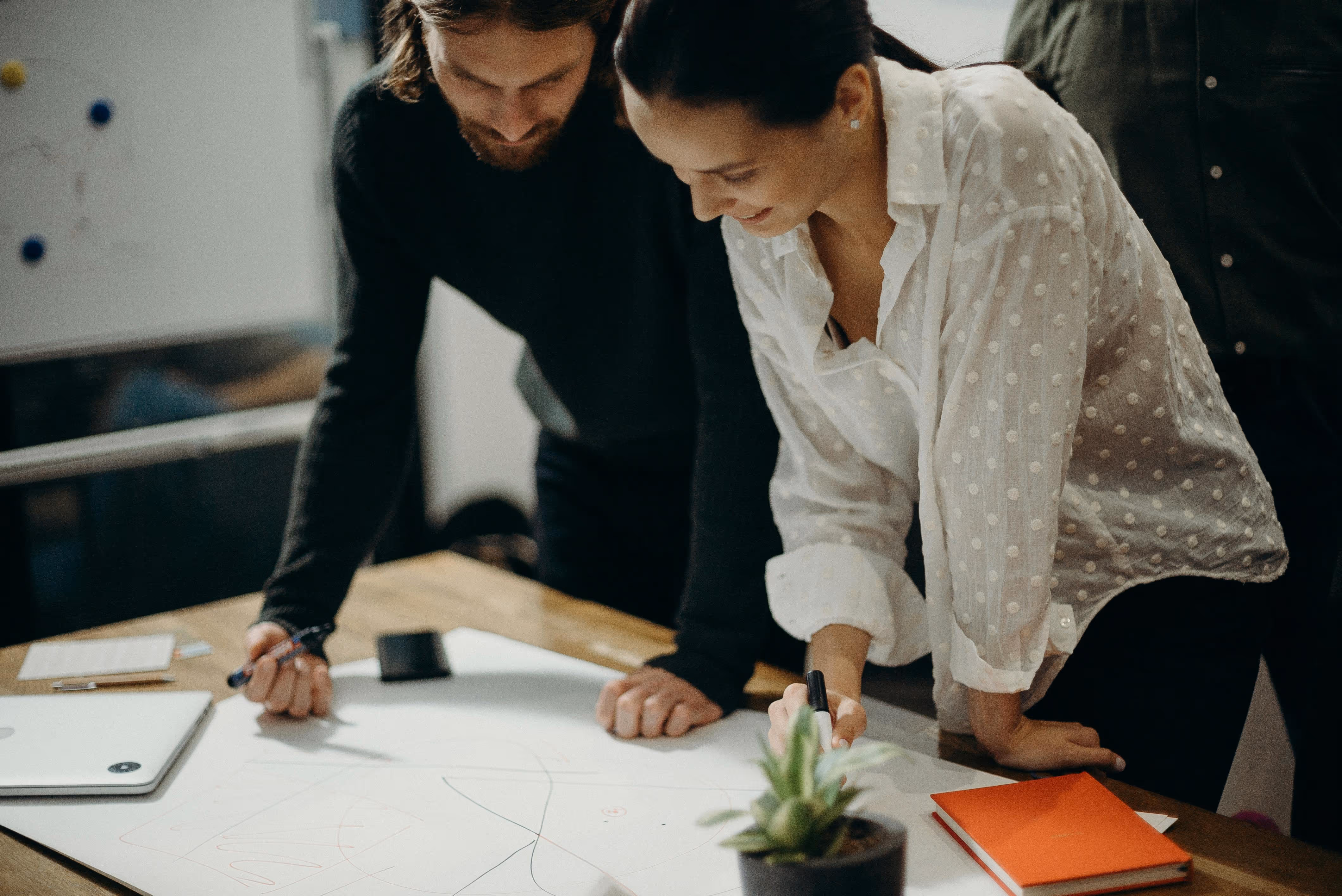 Two colleagues collaborating on a design or brainstorming project, leaning over a whiteboard with markers and notes in a creative workspace