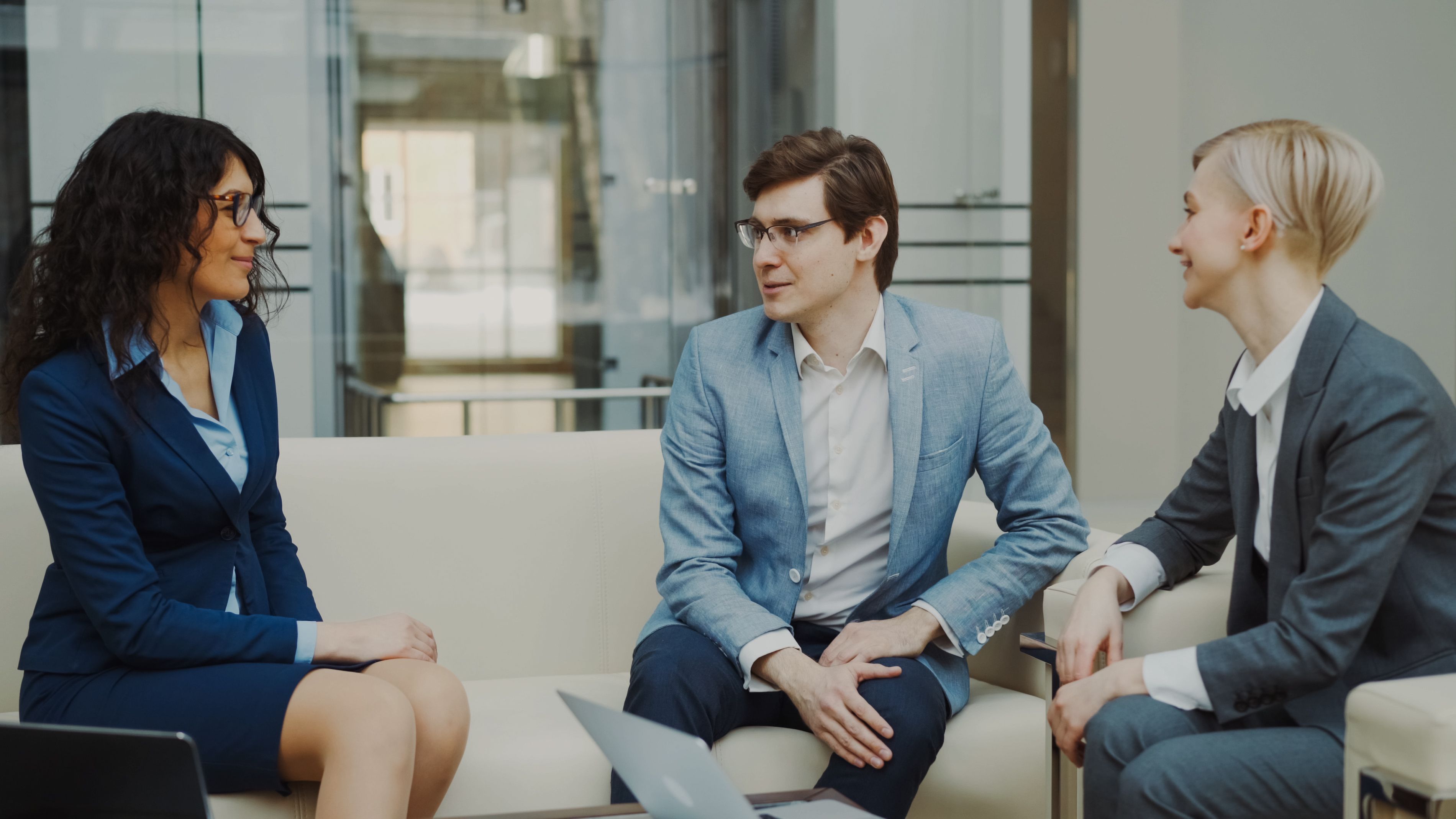 Three professionals having a conversation while sitting on white sofas in a modern office with glass walls, two women and one man all dressed in business attire.