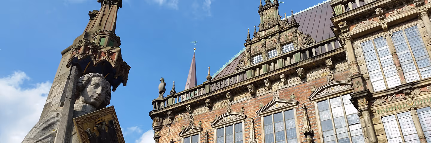 Detailaufnahme der historischen Fassade des Bremer Rathauses mit rotem Backstein, verzierten Fenstern und einer Statue mit Wappen vor blauem Himmel.