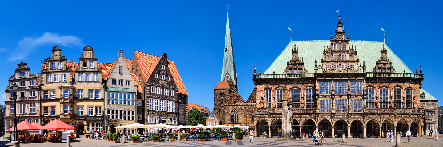 Blick auf den historischen Marktplatz von Bremen mit gotischem Rathaus, Kopfsteinpflaster und Cafés unter Sonnenschirmen bei blauem Himmel.