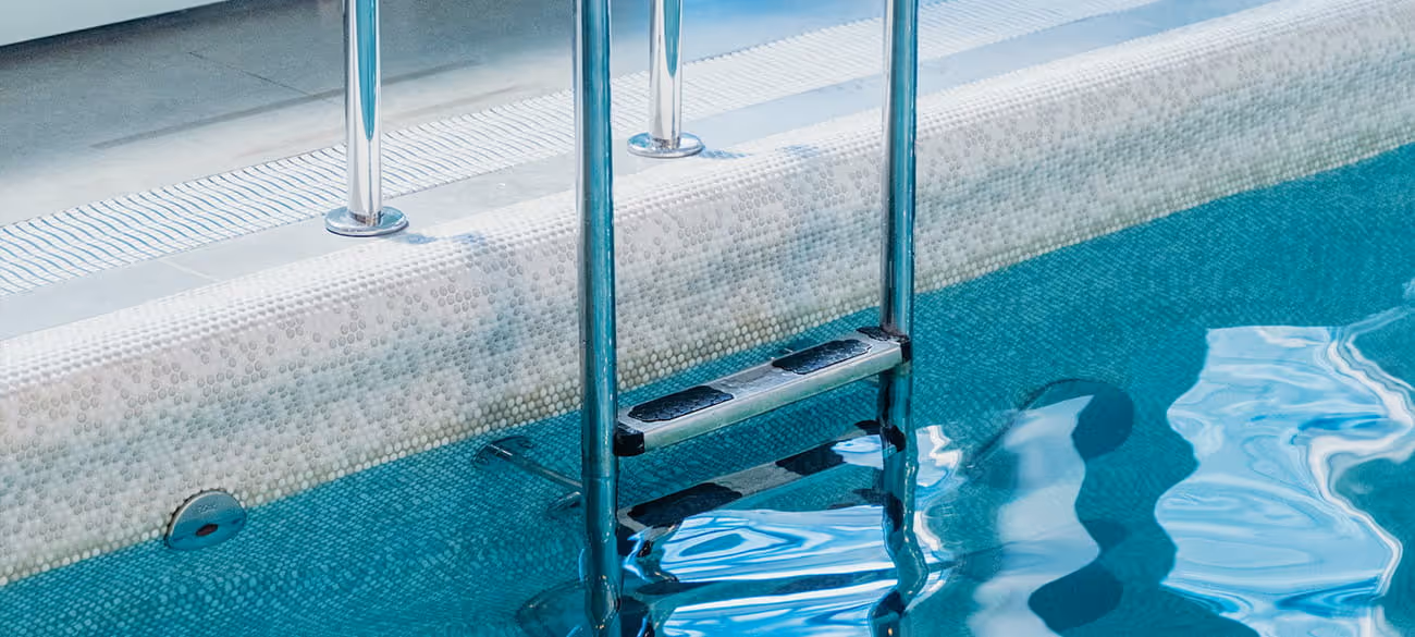 Edelstahlleiter in einem klaren Schwimmbad mit blauem Wasser und beigem Beckenrand.