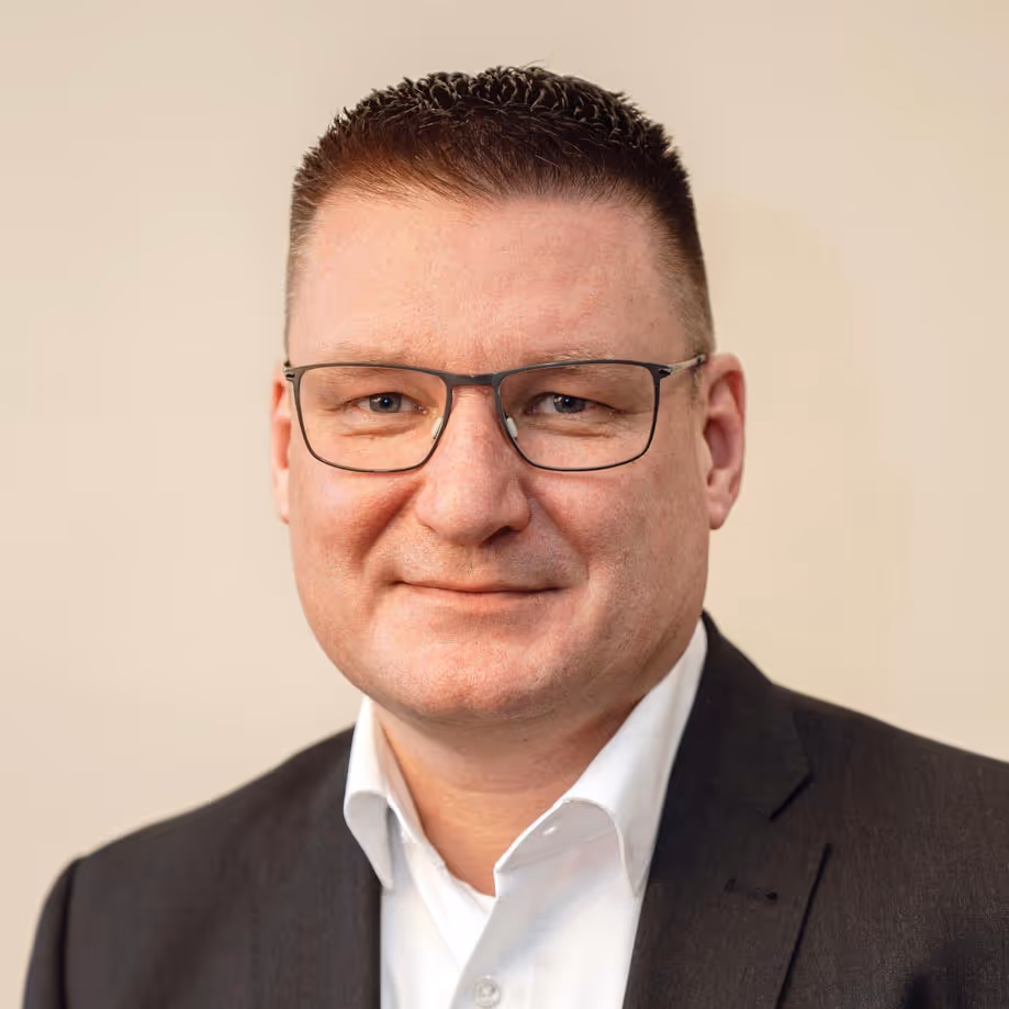 Porträt eines Mannes mit kurzen dunklen Haaren, Brille, weißem Hemd und dunklem Anzug vor beigem Hintergrund.