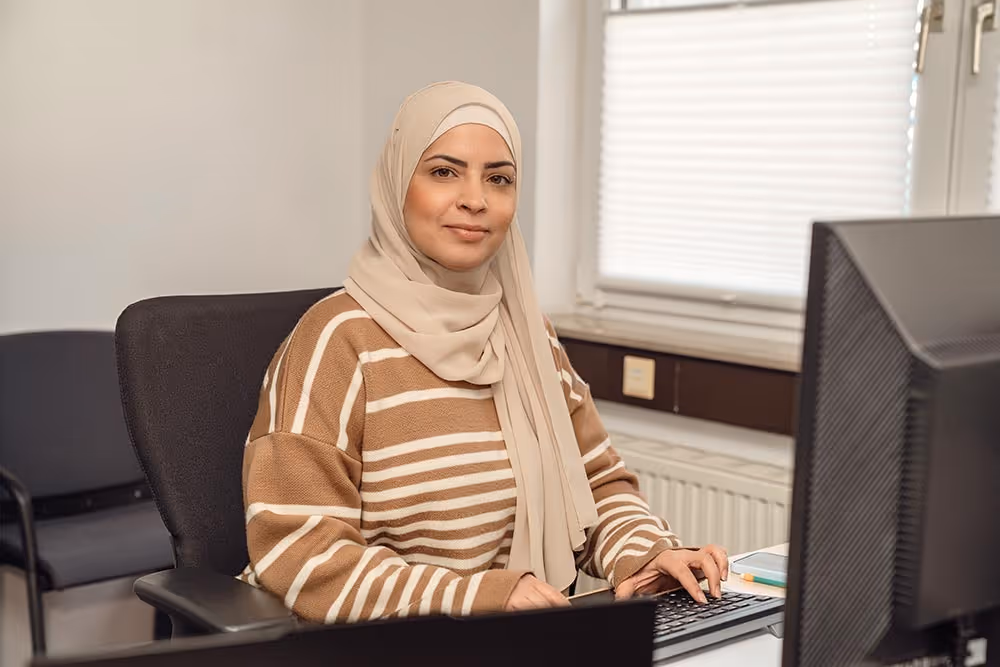 Eine lächelnde Frau mit beigem Hijab und gestreiftem Pullover sitzt an einem Schreibtisch und benutzt eine Tastatur vor einem Monitor.