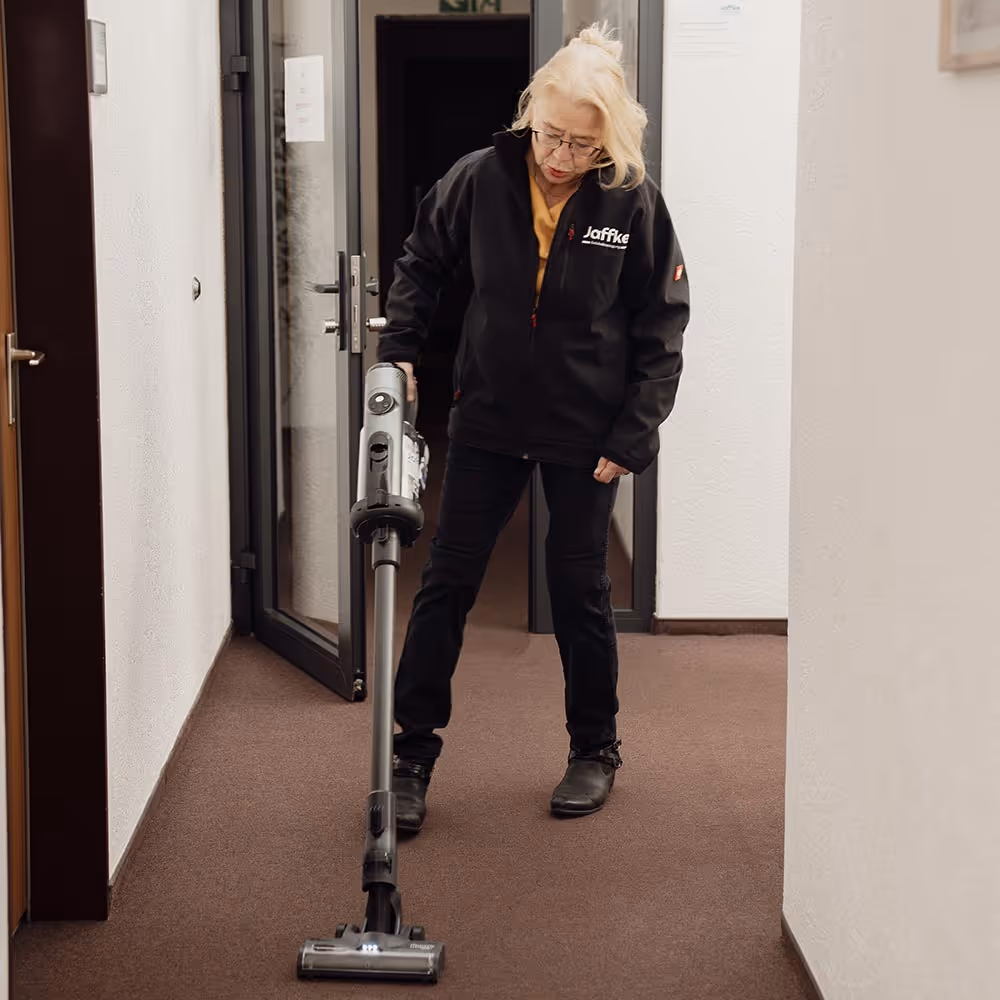 Ältere Frau in Arbeitsjacke saugt einen Flur mit einem Staubsauger.