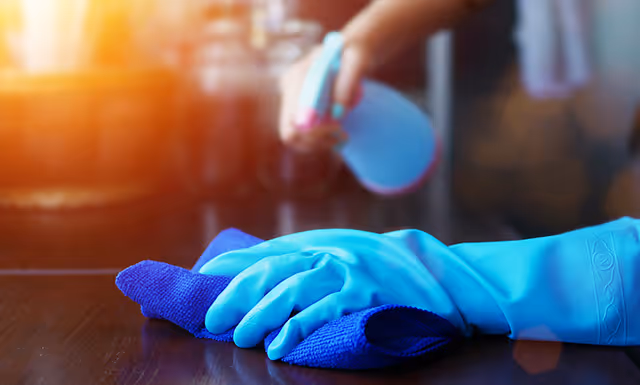 Hand mit blauem Gummihandschuh wischt eine dunkle Tischoberfläche mit einem blauen Tuch.