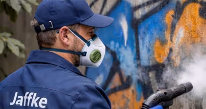 Mann in Schutzmaske und blauem Anzug entfernt Graffiti mit Dampfreiniger an bunter Wand.
