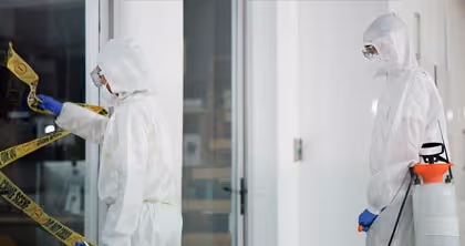 Zwei Personen in Schutzanzügen reinigen einen Tatort mit Warnband und Desinfektionssprühgerät.