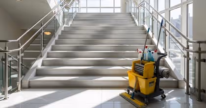 Reinigungswagen mit Putzeimern und Reinigungsmitteln vor einer hellen Treppe mit Edelstahlgeländern.