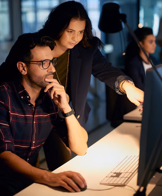 Two engineers reviewing work together on a monitor in a dimly lit office