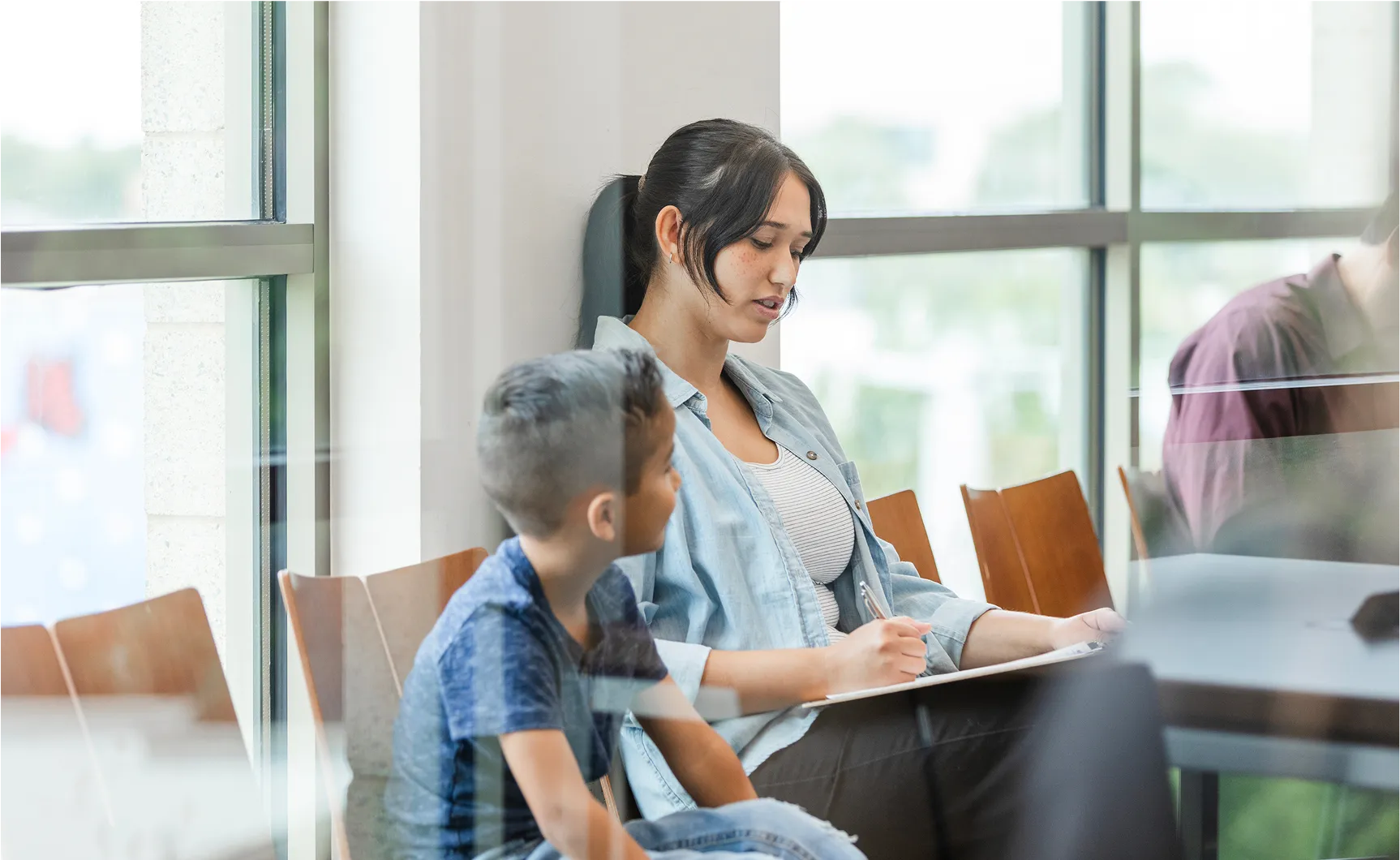 A woman sitting in a modern waiting room with her young son, filling out information on a clipboard.