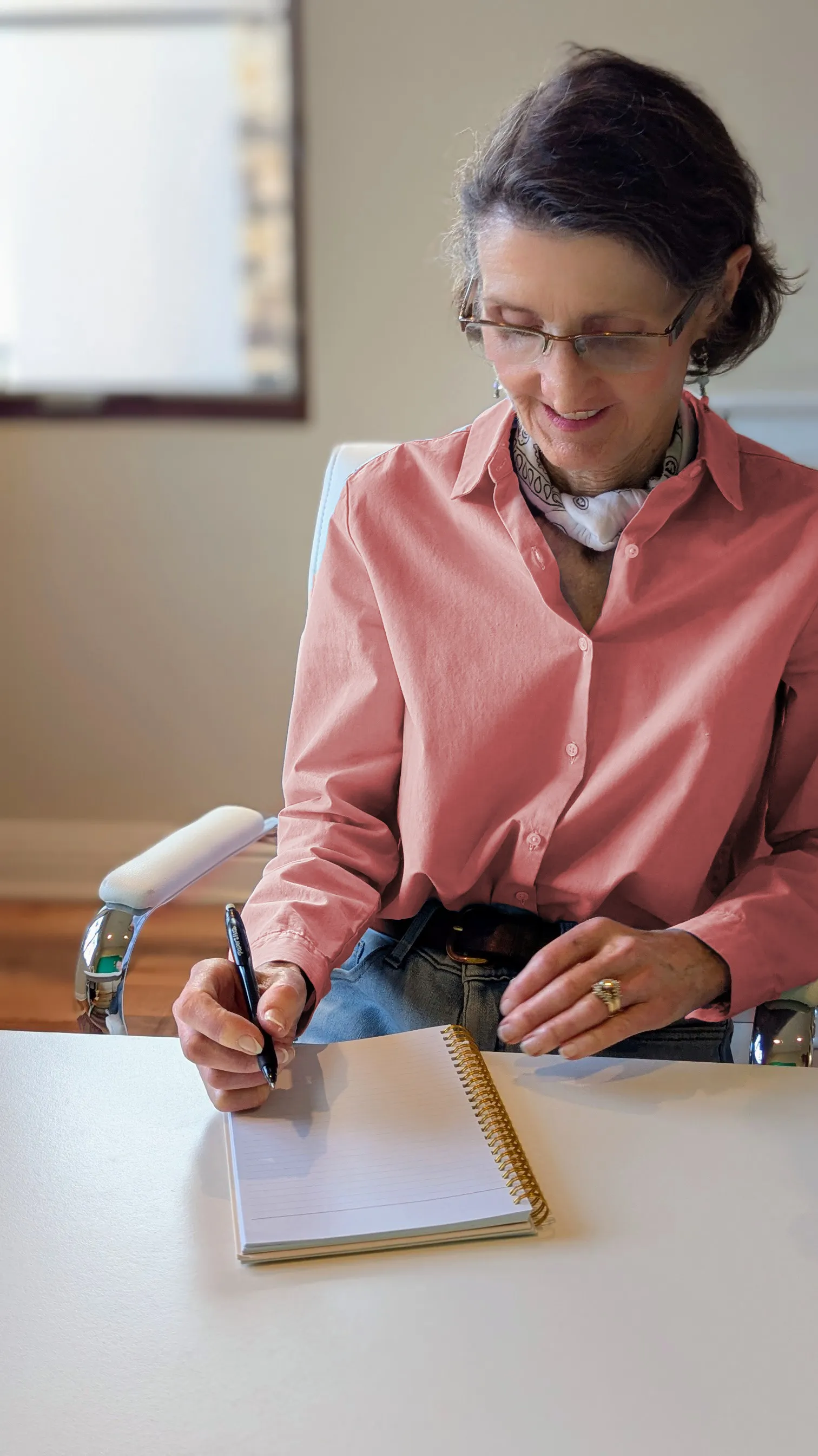 A woman, author Caryn McCann, sits at a desk writing in a notebook, focused on her work.
