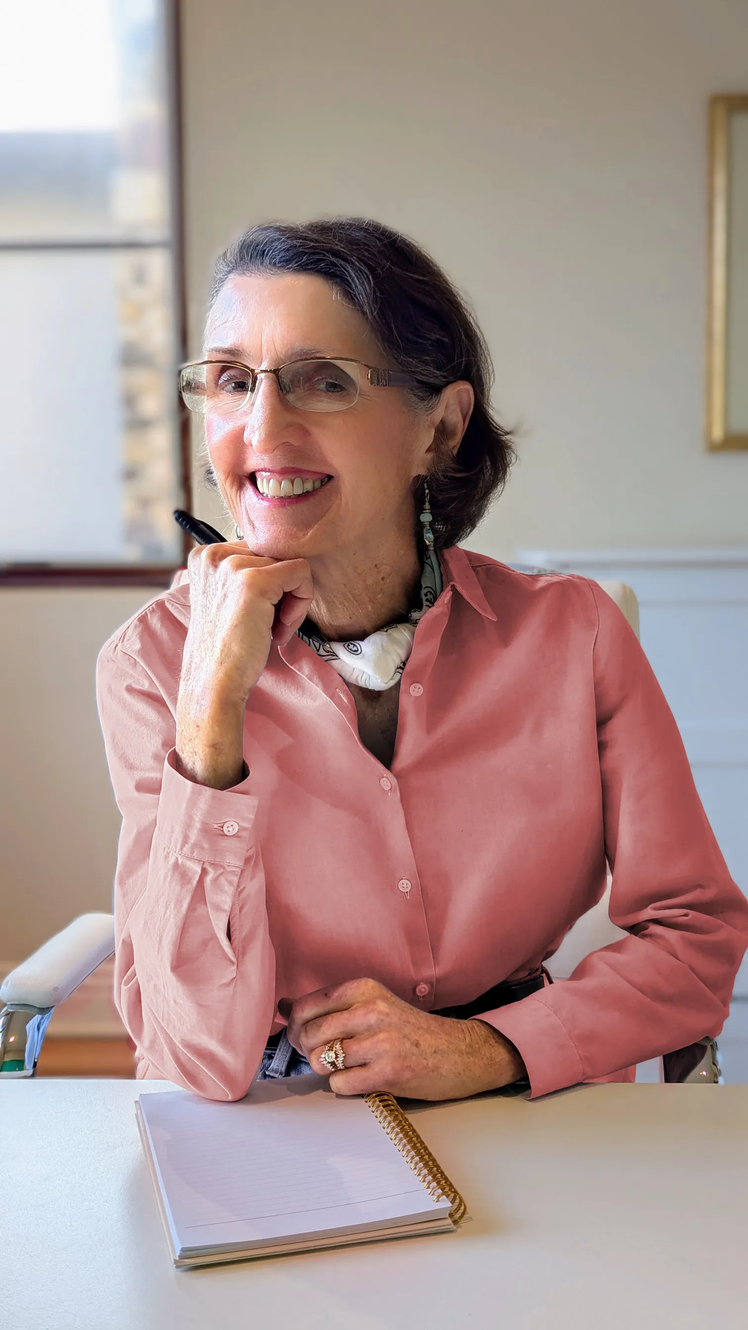 Author Caryn McCann, a woman in a pink shirt and glasses, seated at her desk.