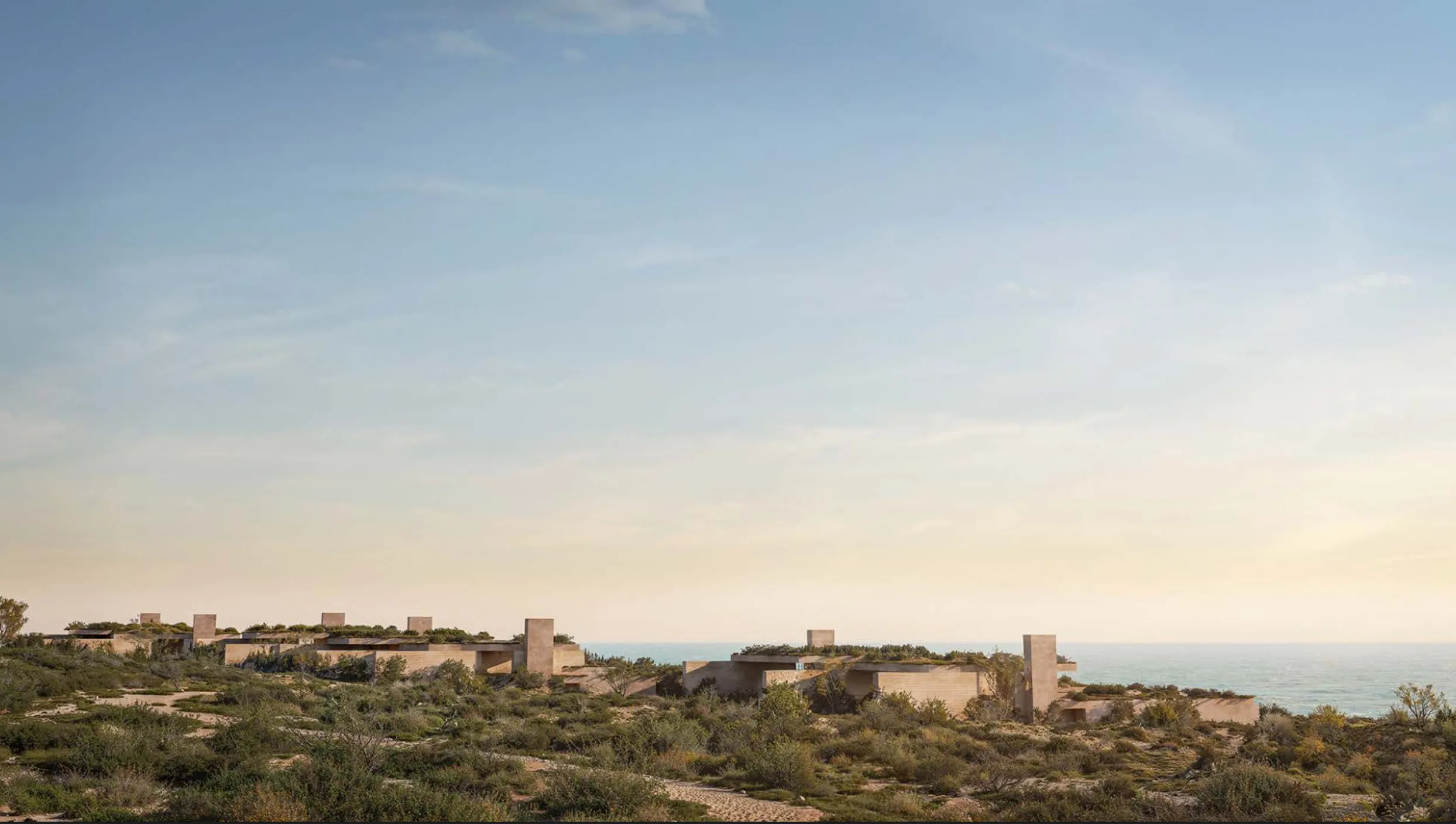 Modern low-profile buildings with green roofs blending into coastal desert landscape overlooking the ocean under a clear sky.