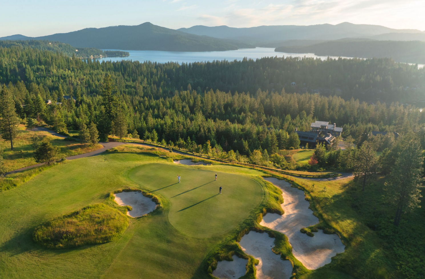 Aerial view of a golf course green with three golfers, surrounded by sand bunkers, forested hills, and a lake in the background during sunset.