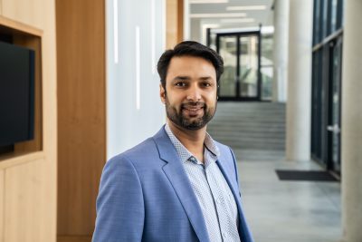 A man with a beard and dark hair wearing a light blue blazer over a patterned button-down shirt, standing in a modern, brightly lit office hallway.