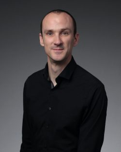 A man with short hair and a slight smile wearing a black button-down shirt, photographed in a professional headshot against a dark grey gradient background.