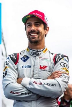 A smiling man with a beard wearing a white Audi e-tron racing suit and a red and white Audi Sport cap, standing with his arms crossed.