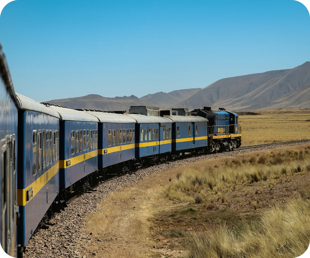 A blue and yellow train travels on a curved track through a vast, arid landscape with distant mountains under a clear blue sky, evoking a sense of adventure.