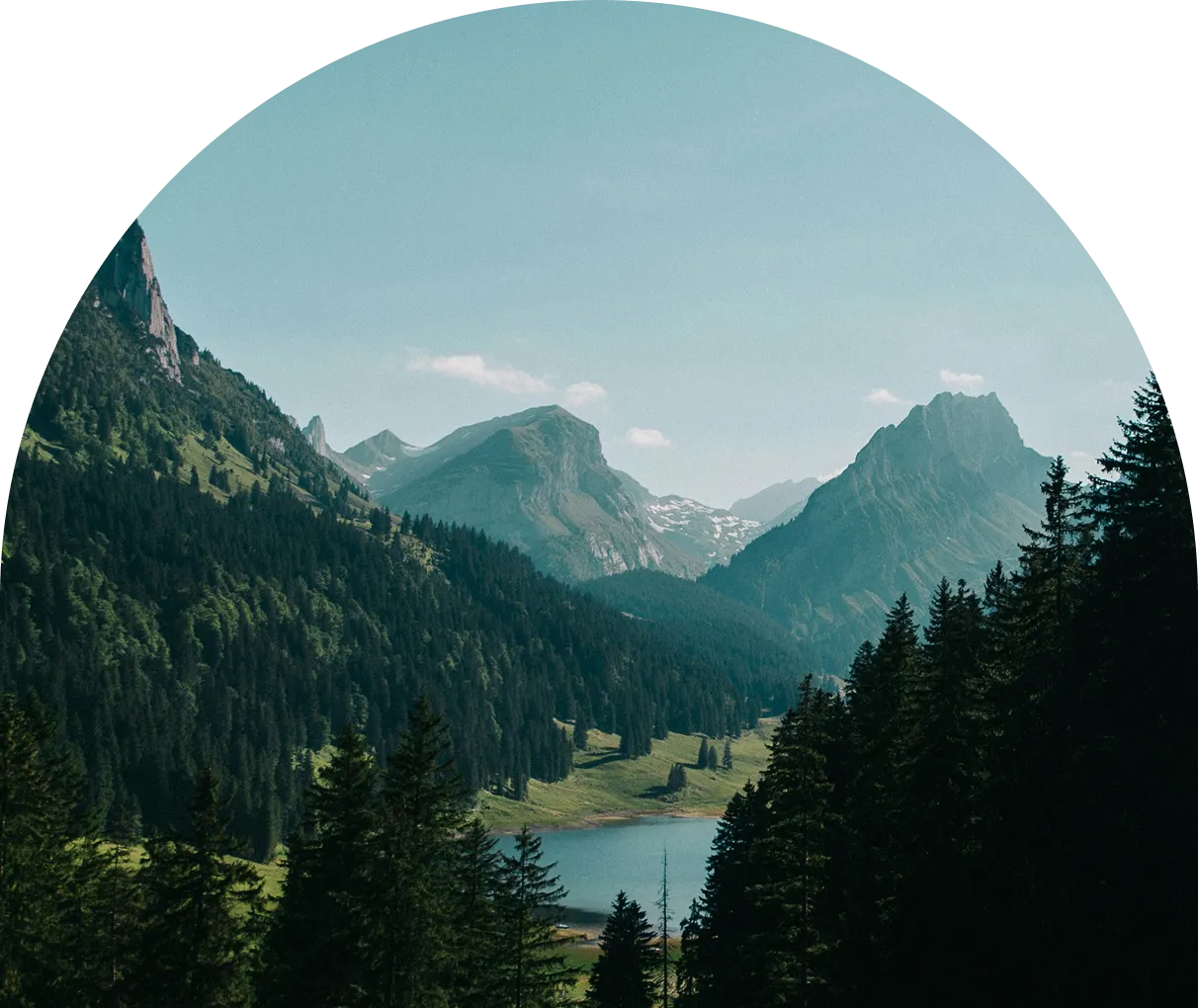 Scenic mountain landscape with snow-capped peaks, dense green forests, and a serene blue lake under a clear sky viewed through a circular frame.