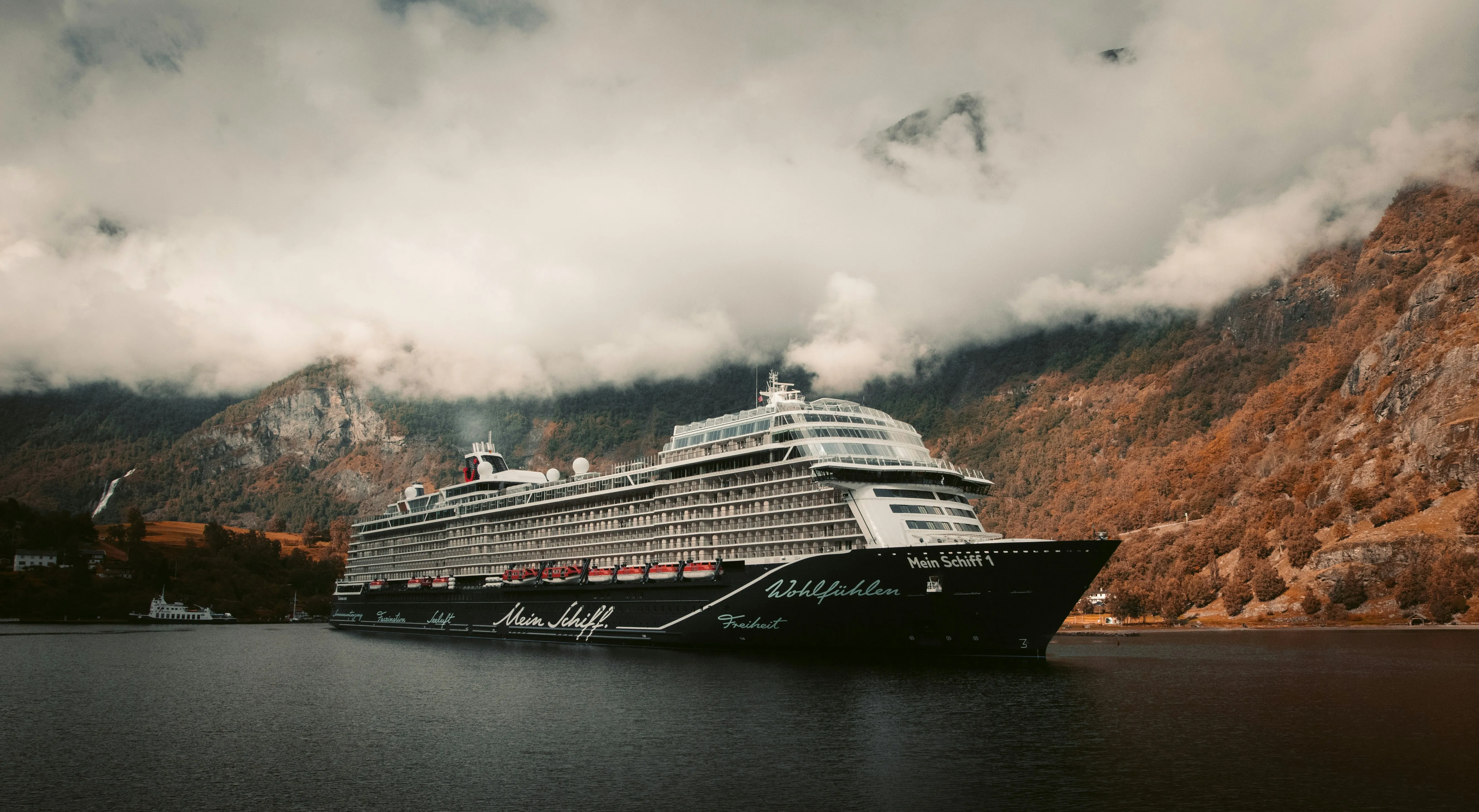 A large cruise ship sails through a serene fjord with misty mountains and autumnal trees in the background. The scene evokes tranquility and grandeur.