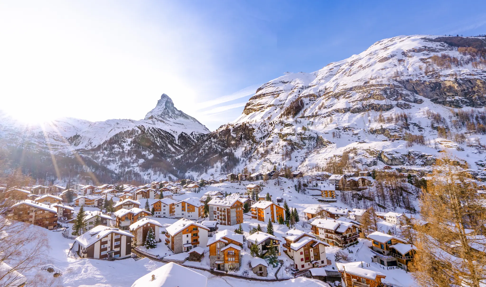 Snow-covered village with wooden houses nestled in the valley, overlooked by majestic, sunlit mountains under a clear blue sky, evoking tranquility.