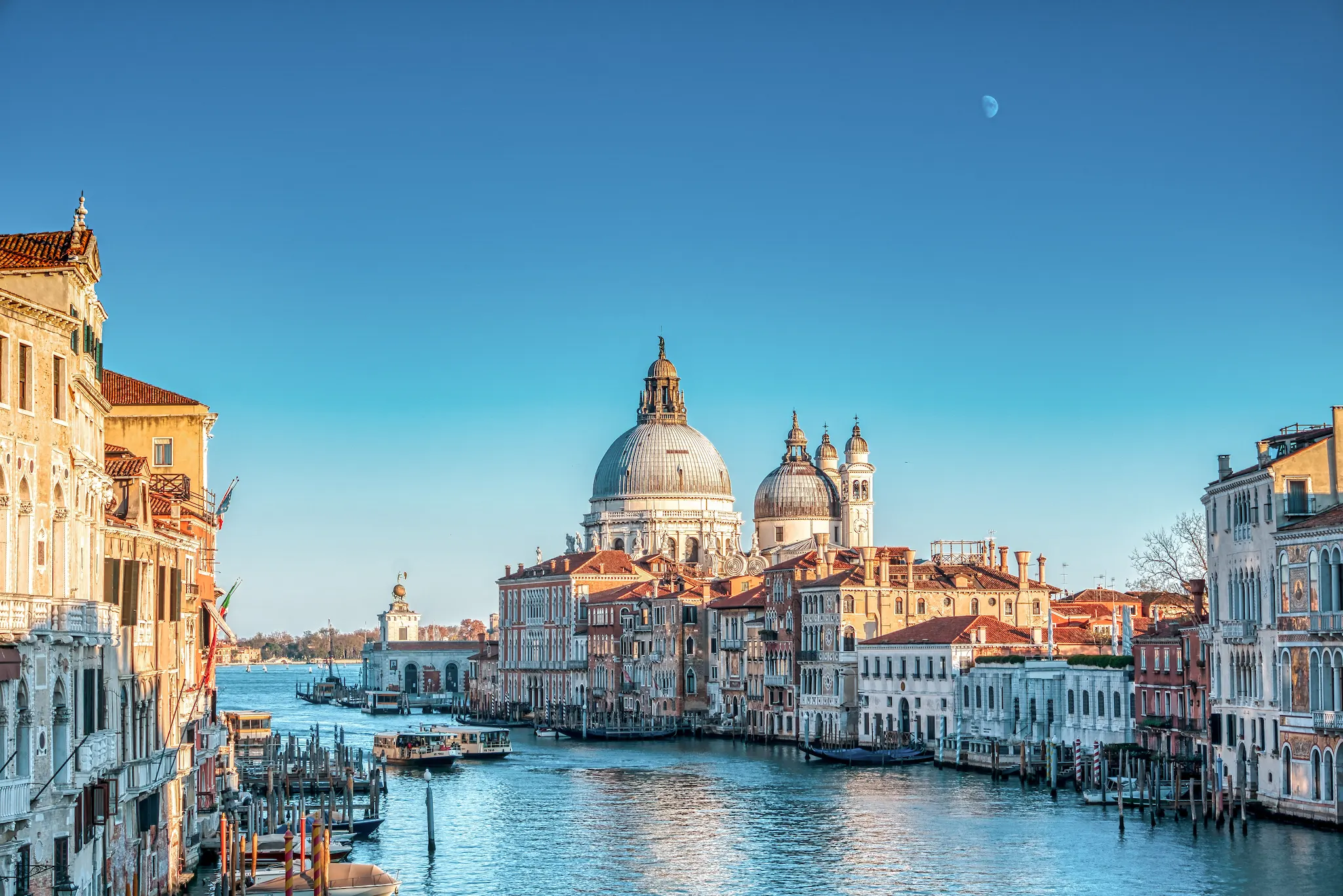 A view of the Grand Canal in Venice, Italy, with historic buildings and the domed Basilica di Santa Maria della Salute under a clear blue sky.