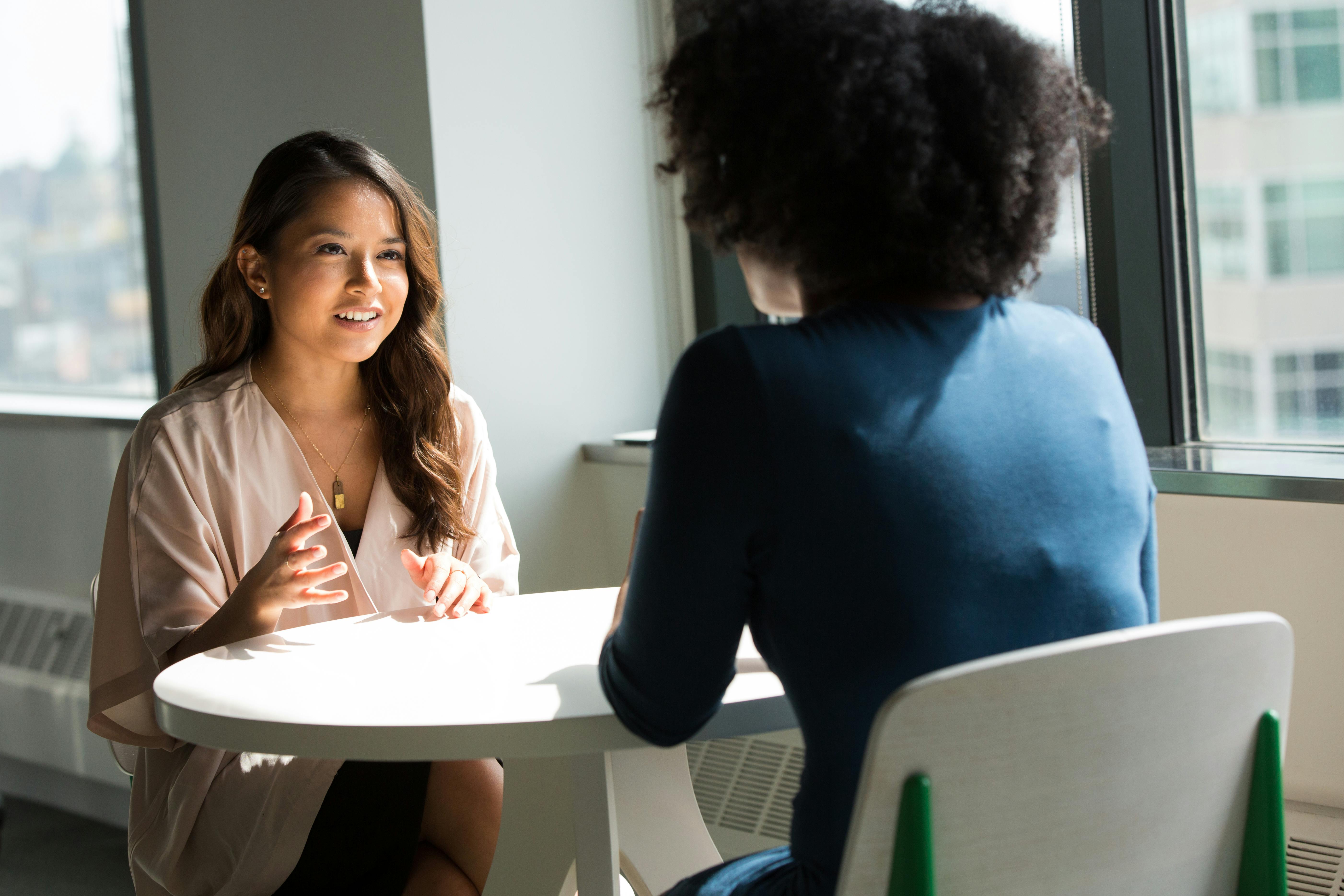 Two women engaged in a professional conversation in a bright, modern office space, representing the collaboration and empowerment of women in business. The interaction highlights the role of women in driving innovative ideas and leadership in the corporate world. 