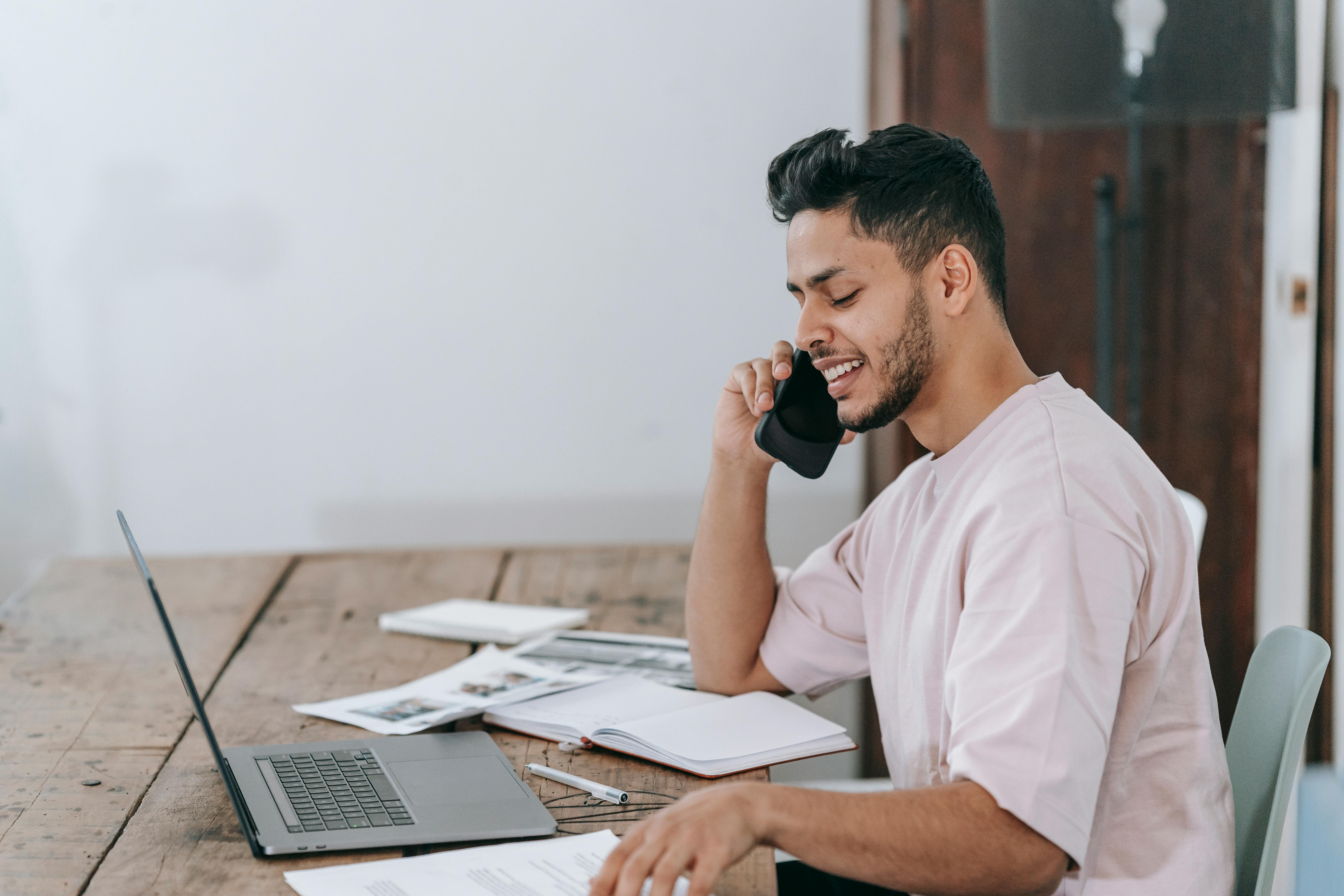 Man smiling while talking on the phone at a desk with a laptop and documents, representing virtual networking and planning for digital marketing meetups and career growth.