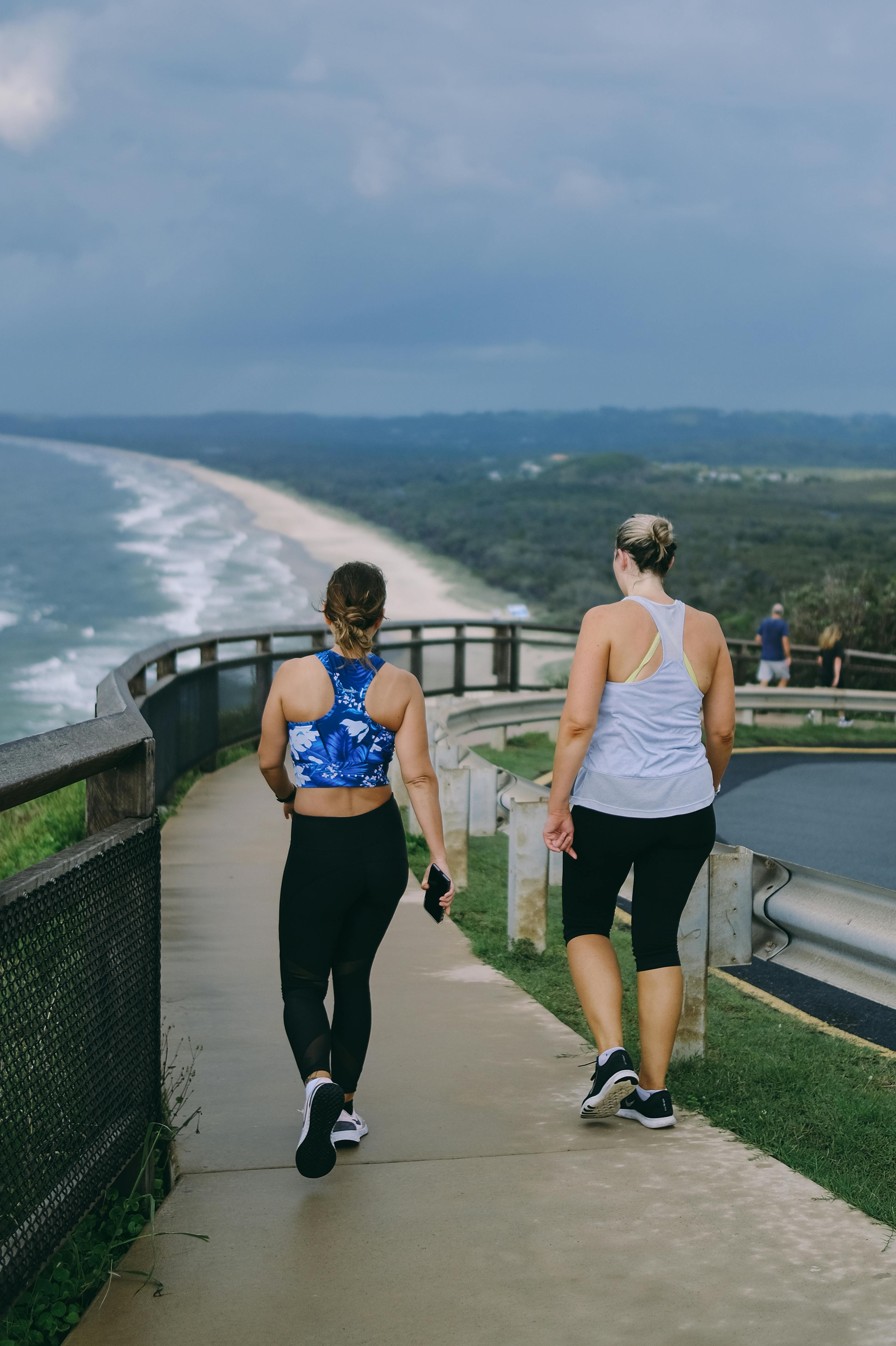 Two women walking along a scenic coastal path in activewear, enjoying exercise and conversation—symbolizing wellness, friendship, and connection in a natural setting.