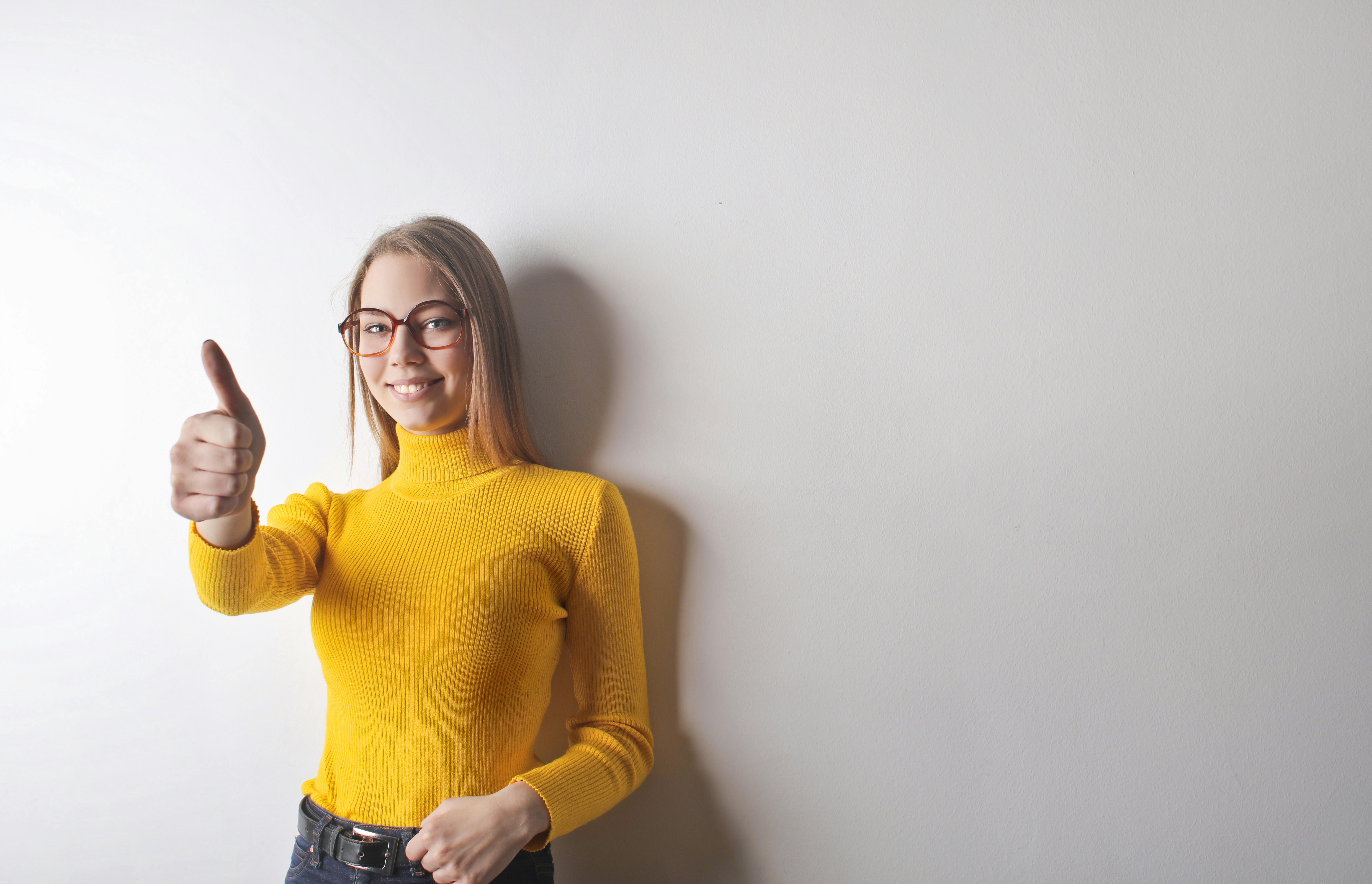 Confident young woman in a yellow jumper giving a thumbs-up, symbolising self-belief, social growth, and breaking social barriers through new activities.