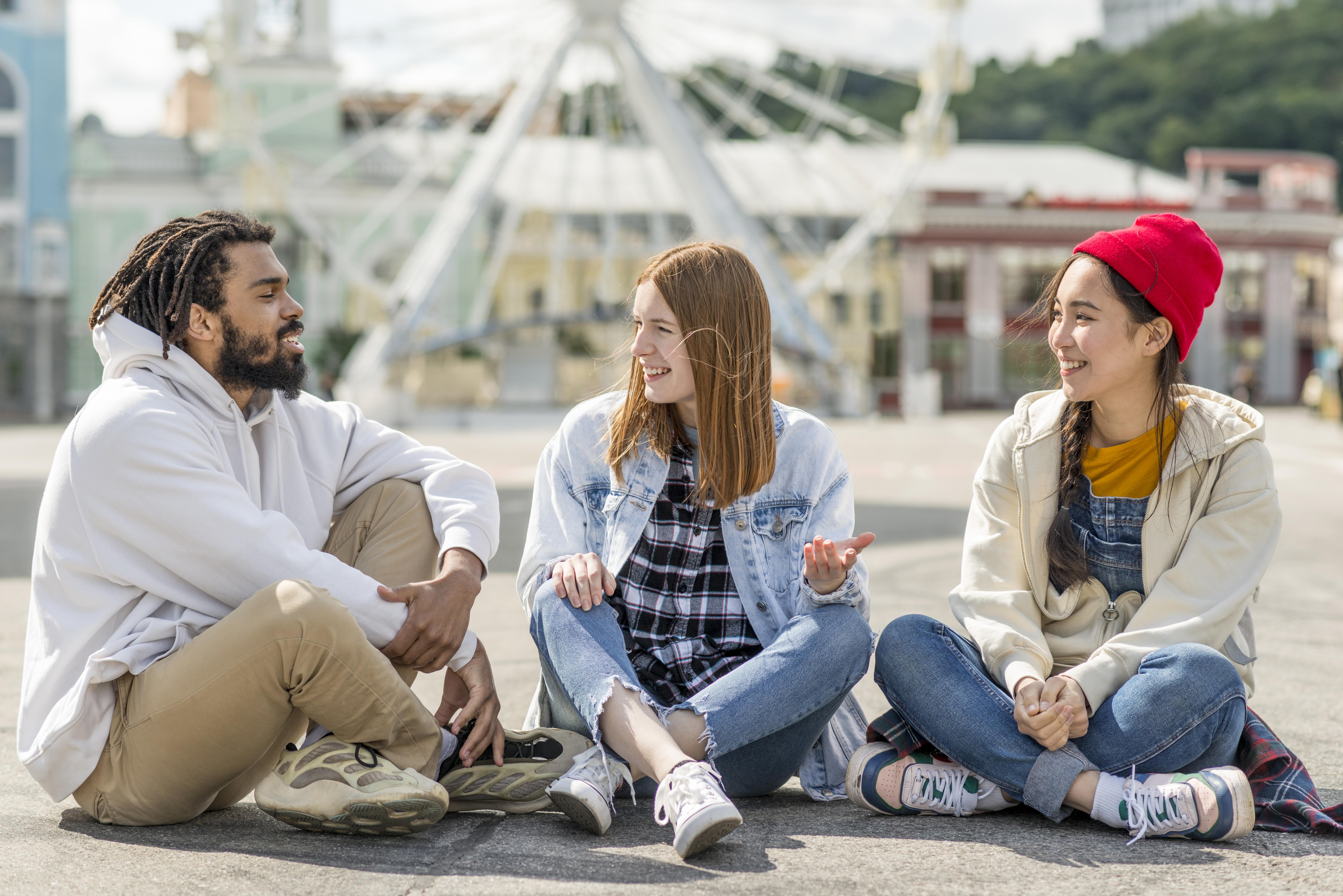 Three young adults sitting on the ground near an urban ferris wheel, talking casually and smiling, illustrating relaxed modern adult friendships and low-pressure connection in Australia.