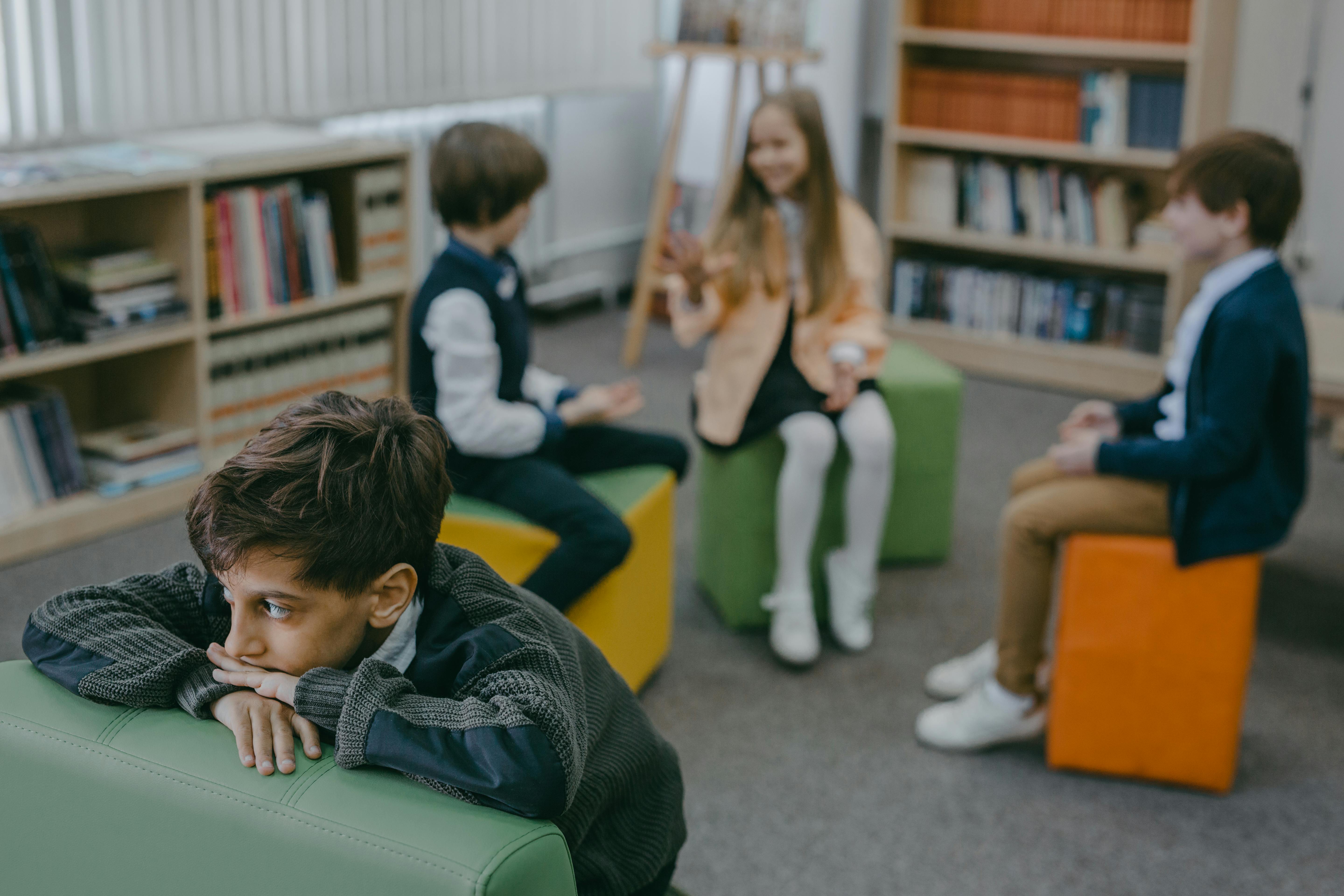 Young boy sitting alone on a green seat in a school library while three other children talk together in the background, illustrating childhood loneliness and social exclusion.