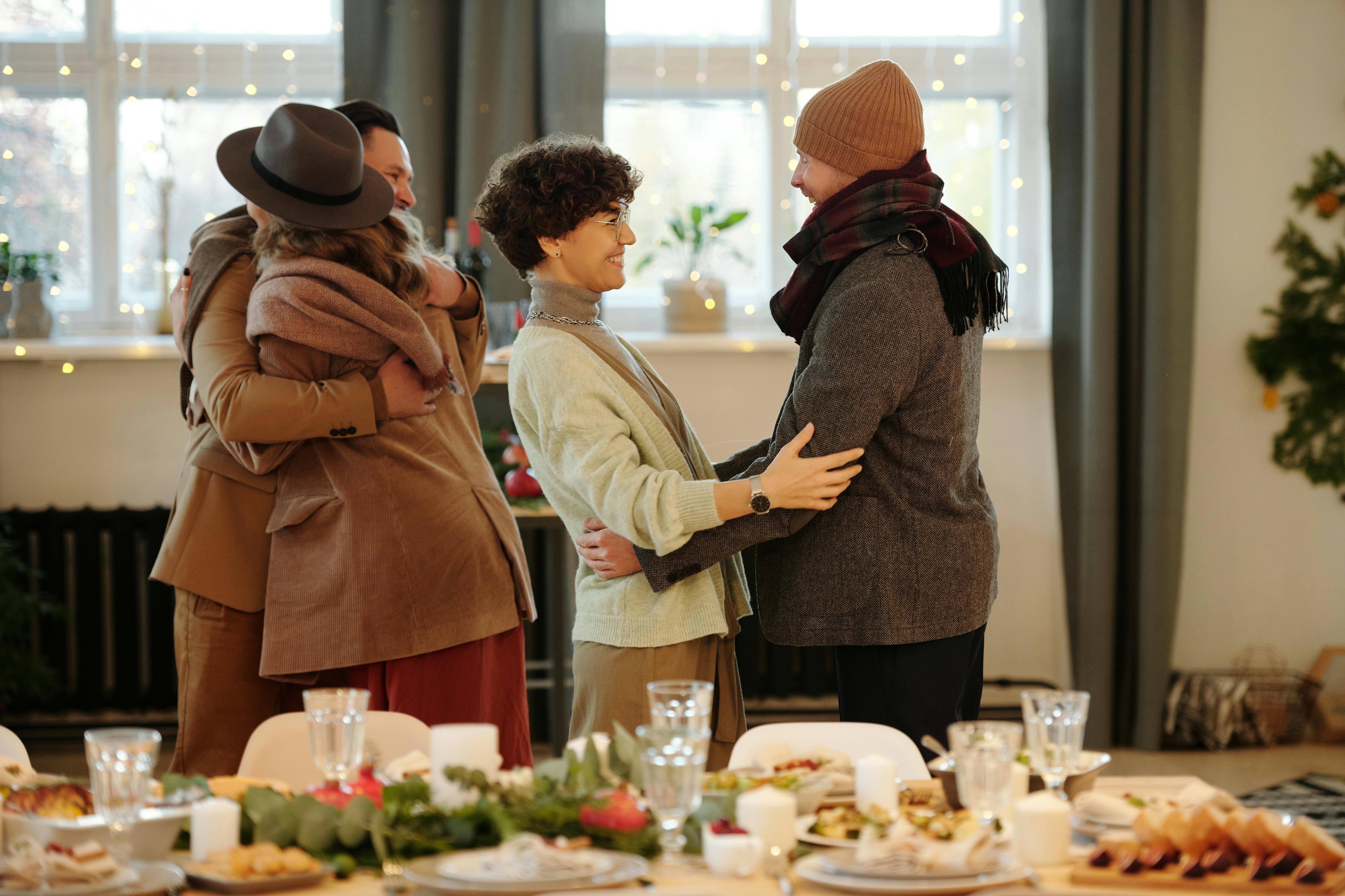 Two pairs of adults greeting and hugging in a warmly lit home, standing near a festive dinner table, illustrating parents building friendships and support circles through everyday social gatherings in Australia.