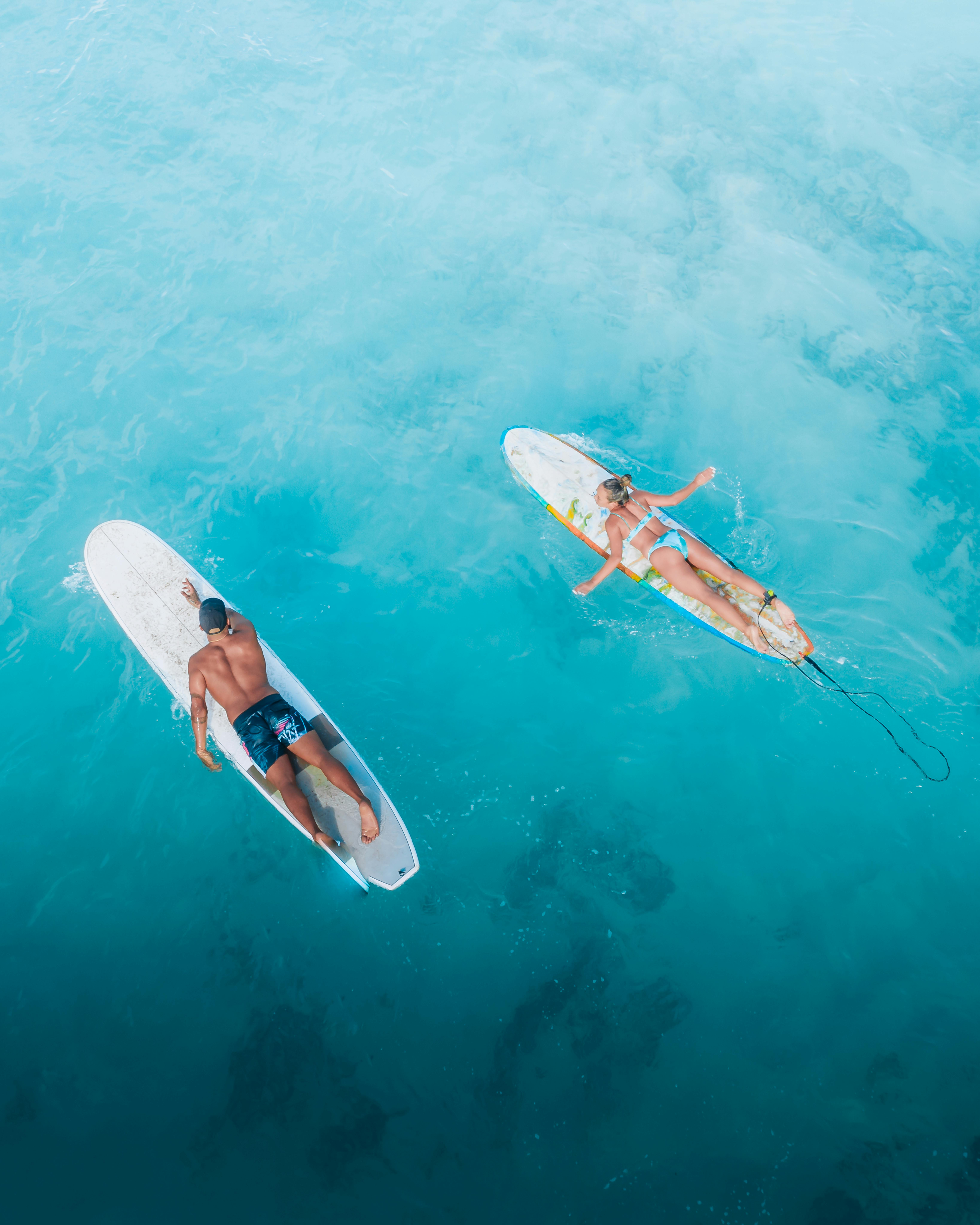 Two people lying on surfboards in clear blue ocean water, paddling side by side, representing how shared hobbies and relaxed outdoor activities in Australia can help build real social connections without awkward big events.