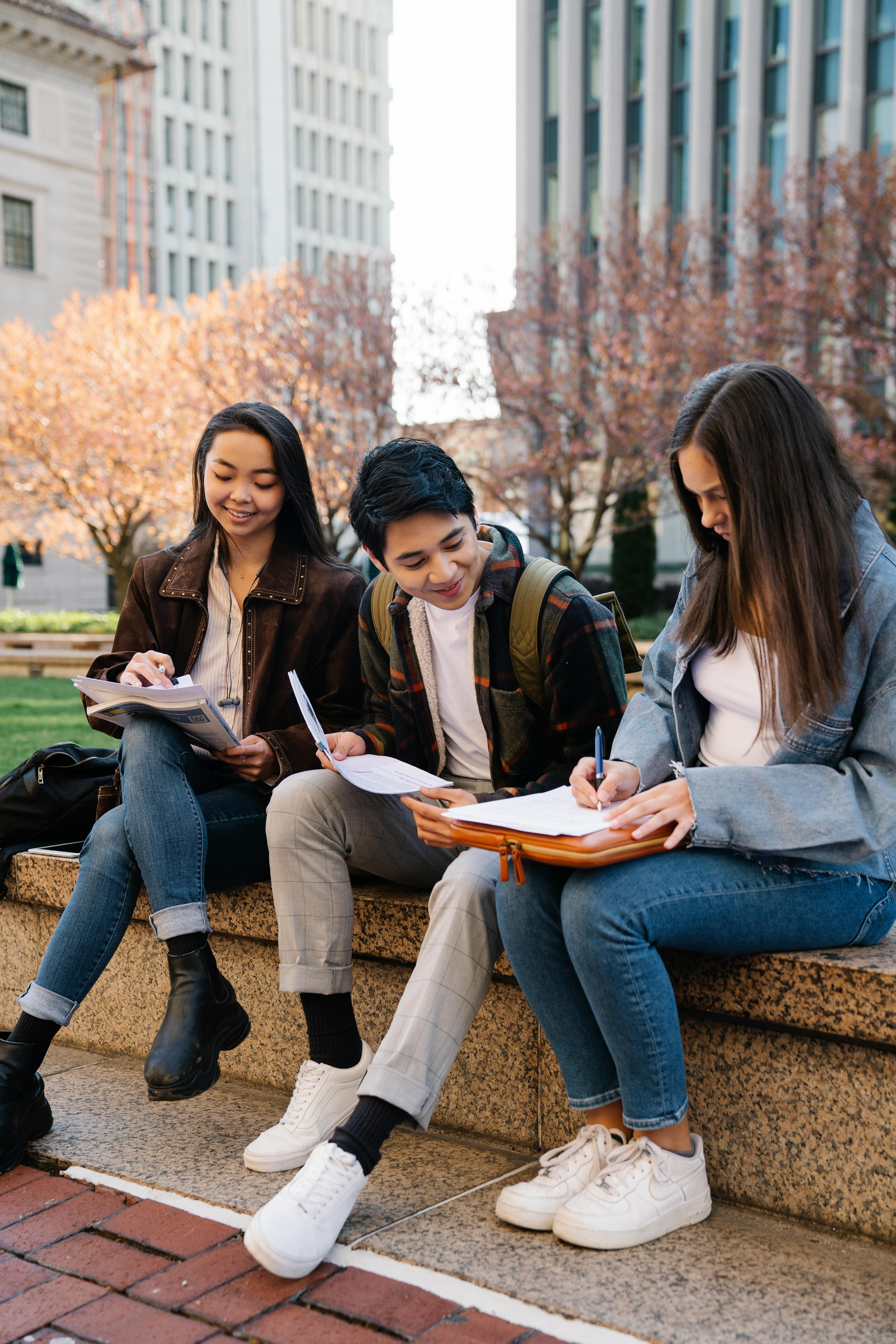 Three university students sitting on a campus bench with notebooks, studying and talking together, representing how everyday conversations, classes, and small study catchups can grow into genuine friendships at uni in Australia without pressure or awkward expectations.