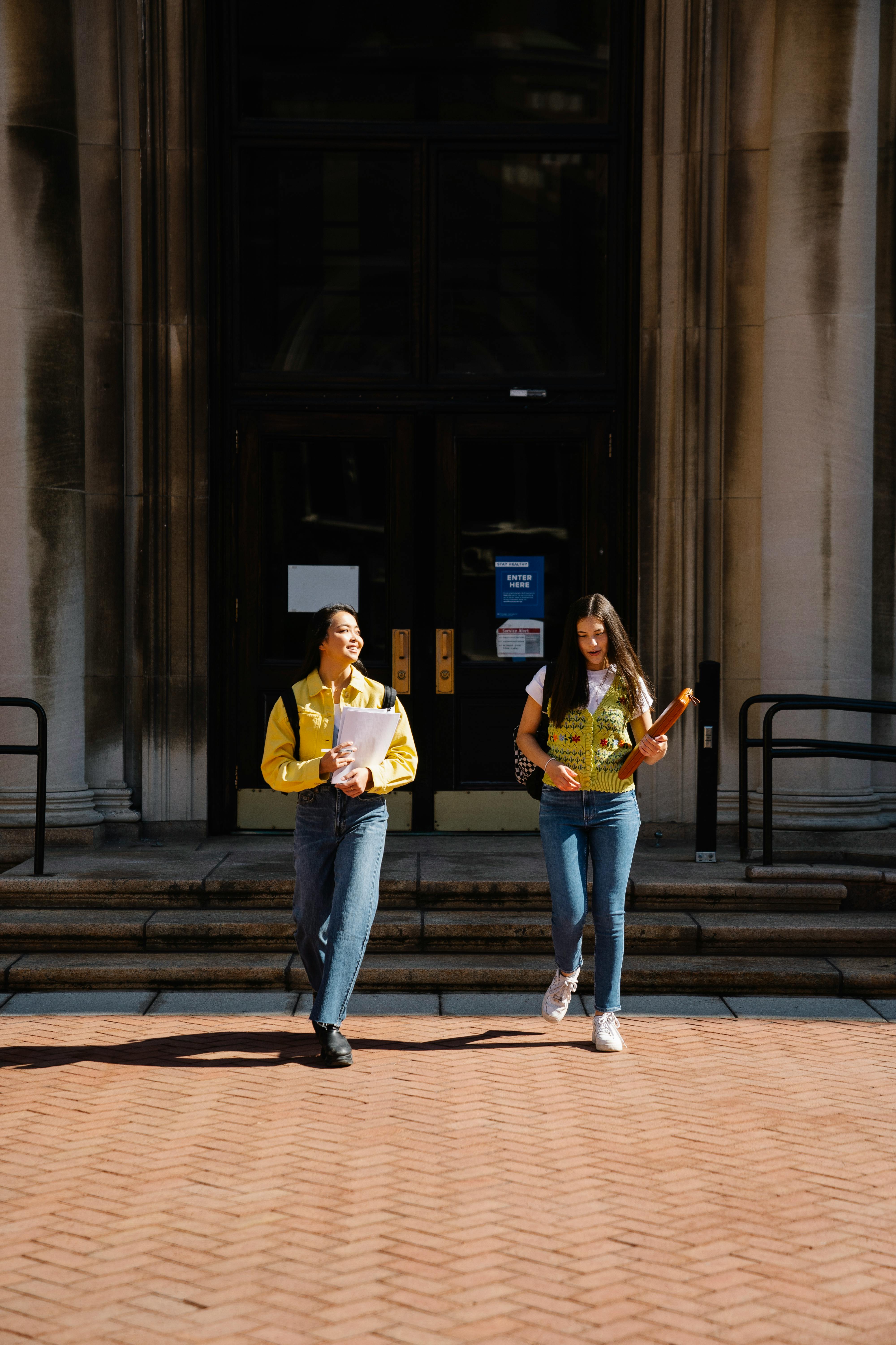 Two international students walking out of a university building, talking and smiling with books in hand, representing how everyday campus moments and relaxed conversations can grow into real friendships in Australia without pressure or pretending.