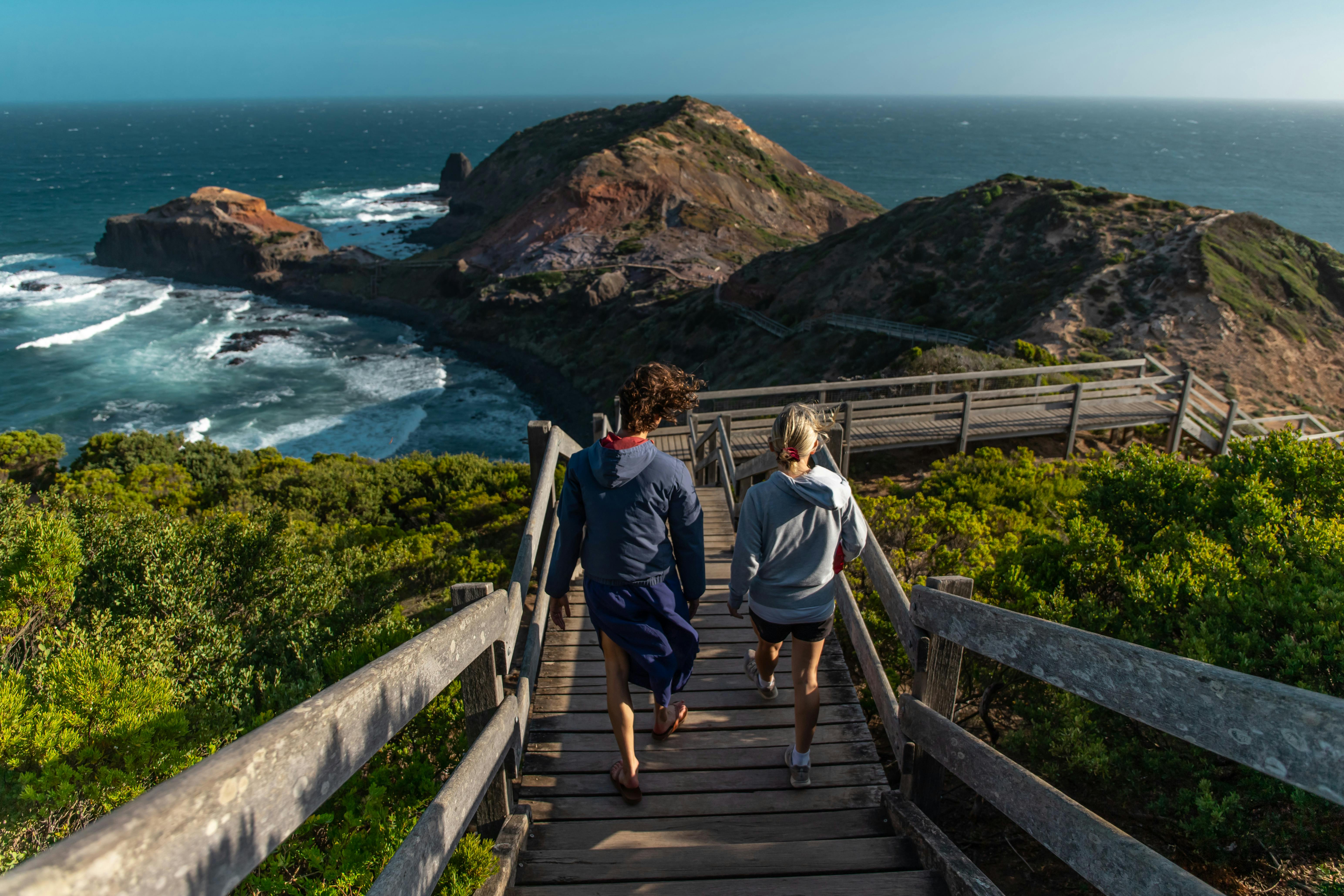Two friends walking down a coastal boardwalk overlooking the ocean, representing how shared outdoor adventures and relaxed one-on-one hikes in Australia can help you safely find the right hiking buddy without big, awkward groups.