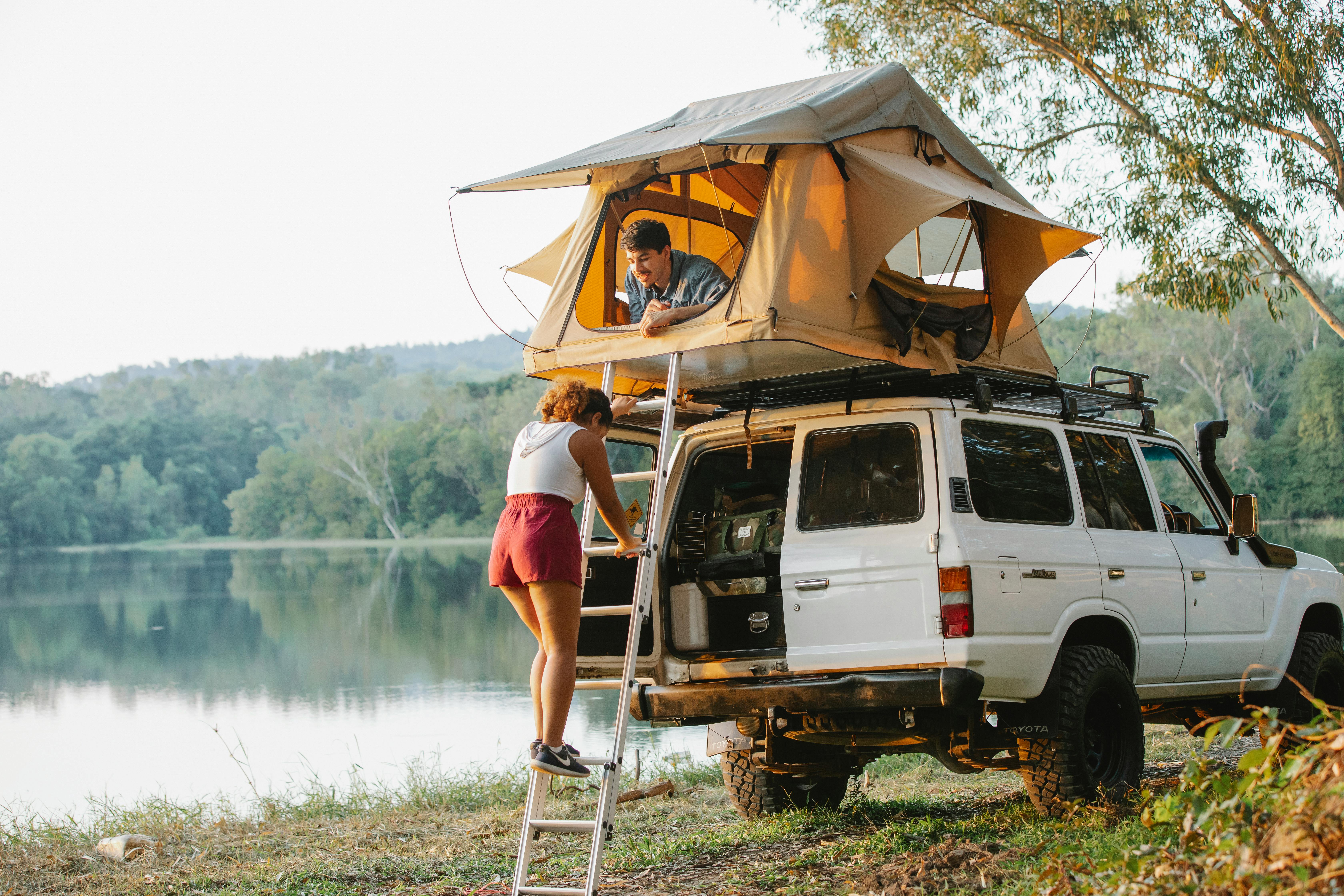 Two friends camping beside a lake with a rooftop tent on a 4WD, one climbing the ladder and one relaxing above, representing safe, friendly camping buddies in Australia and how the right match can make trips feel relaxed, fun, and secure.