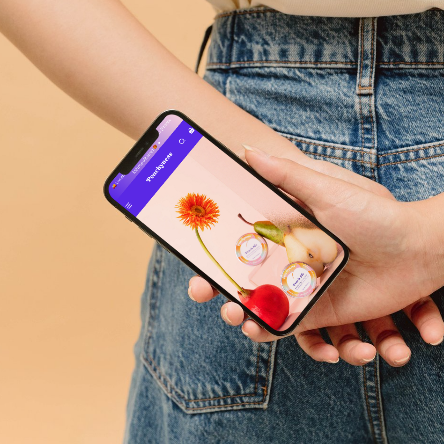 Person holding a phone displaying the Peachyness website with images of a flower, pear, and skincare products.