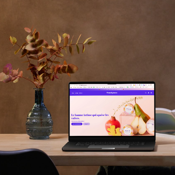Laptop on wooden desk displaying Peachyness website with intimate balm products, next to a decorative vase with autumn-colored eucalyptus branches.
