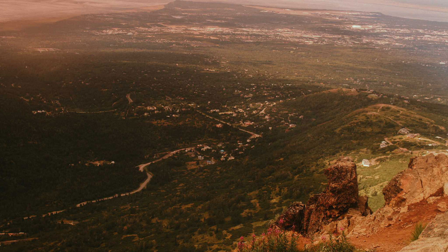 A scenic photograph of the view from the top of some cliffs overlooking a town interspersed with trees.
