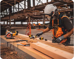 Construction worker in safety gear using power tools to measure and cut timber on-site — part of a Diaz Murphy Studio build