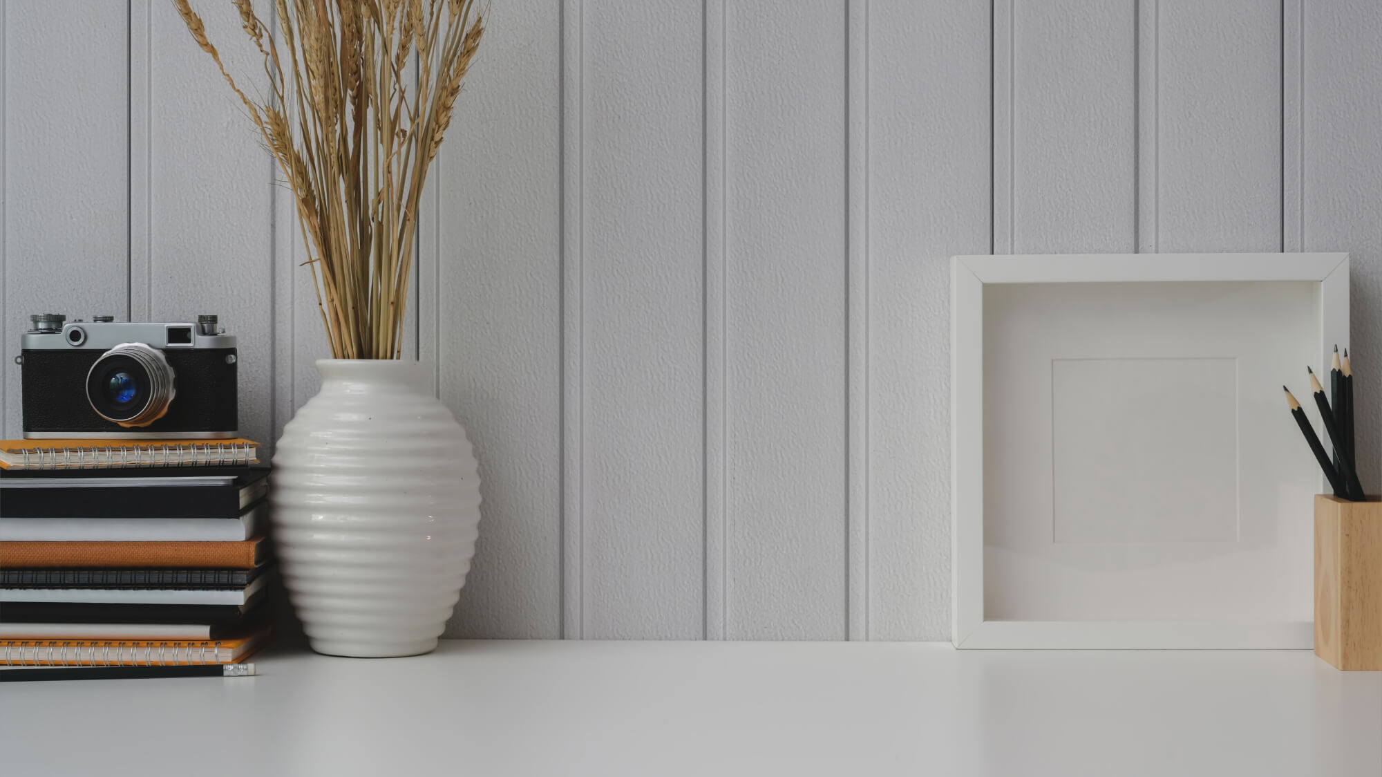 Stack of notebooks with a vintage camera on top beside a white ceramic vase with dried wheat stalks, next to a blank white picture frame and a wooden holder with black pencils on a white desk against a light gray paneled wall.