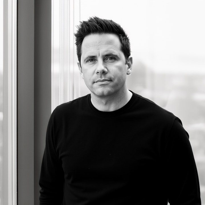 Black and white portrait of a man with short dark hair wearing a black shirt standing by a window.