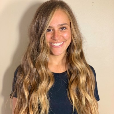 Smiling woman with long, wavy blonde hair wearing a black shirt standing against a plain light-colored wall.