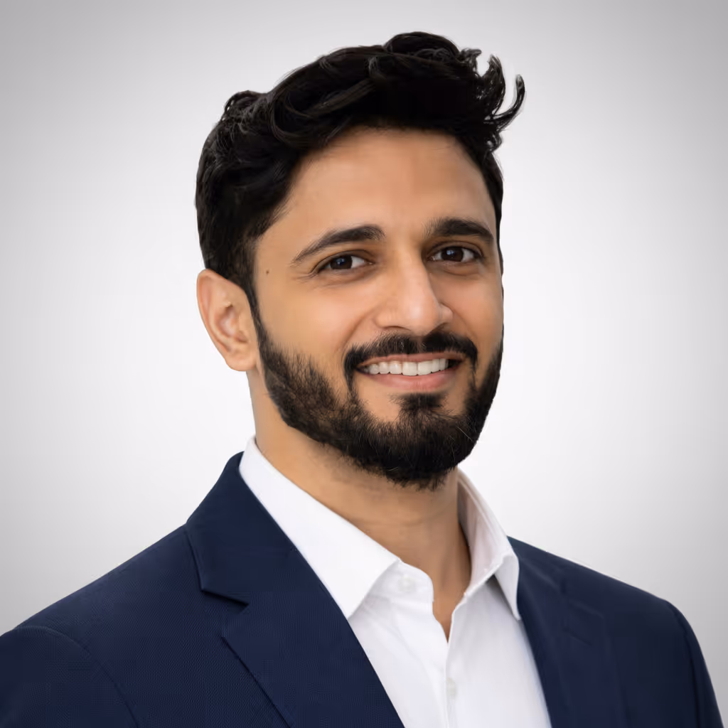 Smiling man with beard and dark hair wearing a navy blue suit jacket and white shirt against a light gray background.