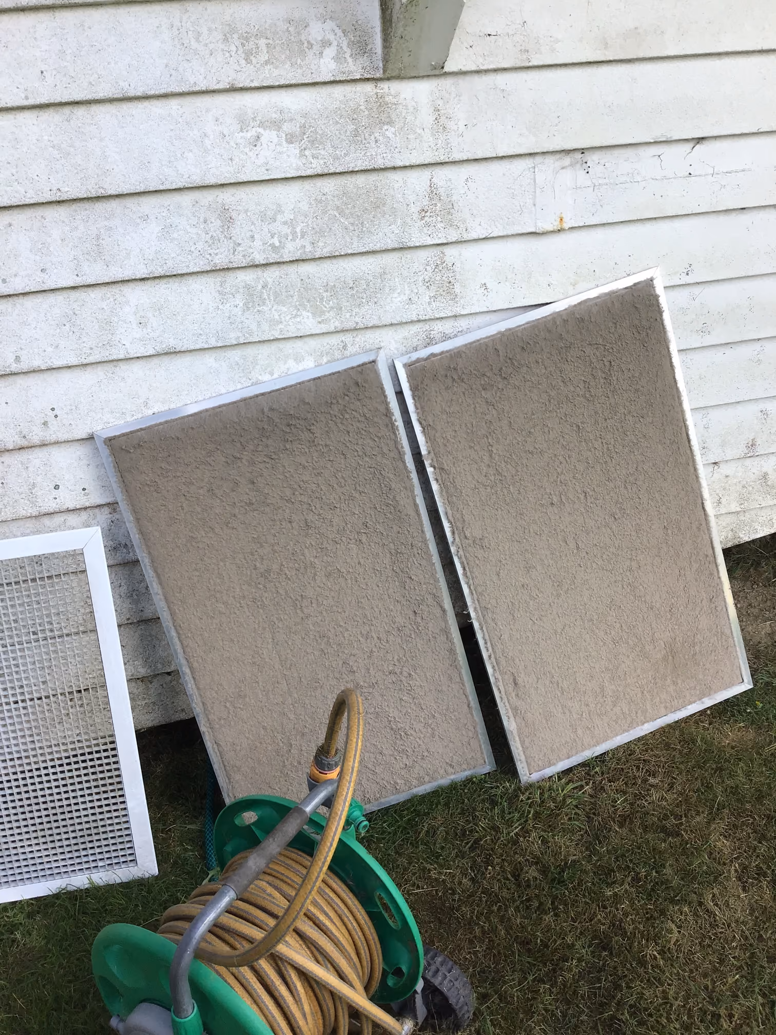 Two heavily dust-covered air filters leaning against the exterior wall of a white house, next to a garden hose reel with a coiled yellow hose on green wheels, resting on grass.