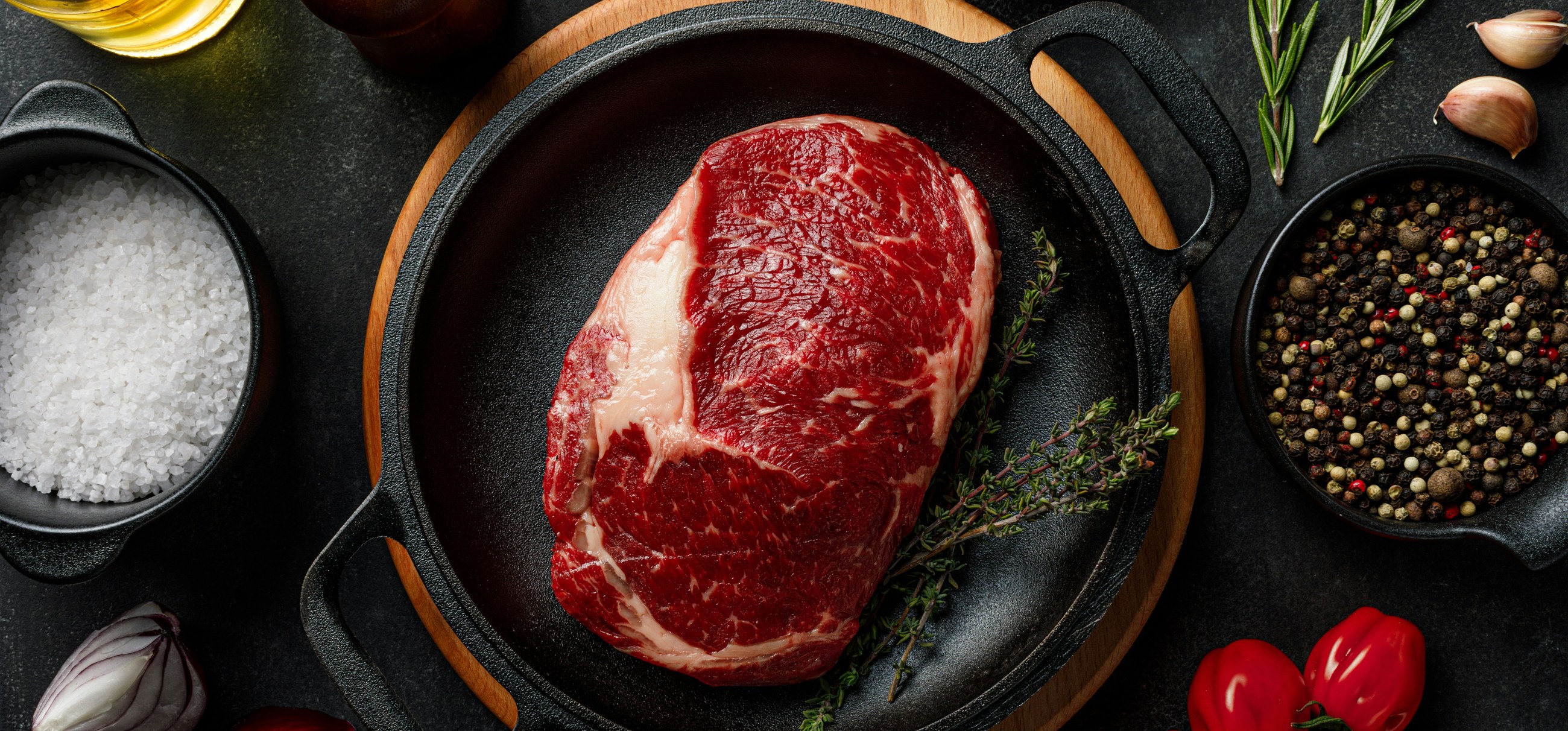Ein rohes Ribeye-Steak in einer Gusseisenpfanne mit Salz, Pfefferkörnern und Kräutern