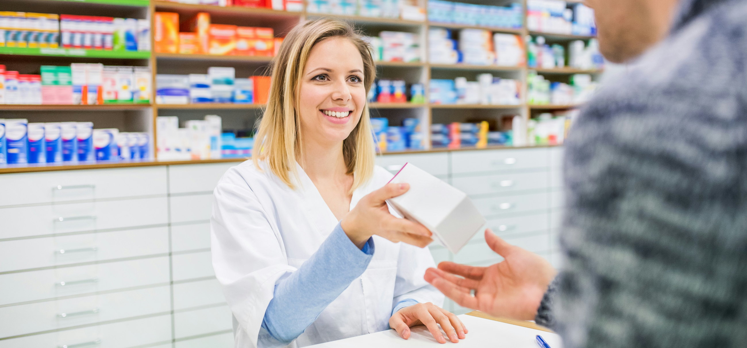 A pharmacist hands a medicine box to a customer across the counter of a community pharmacy